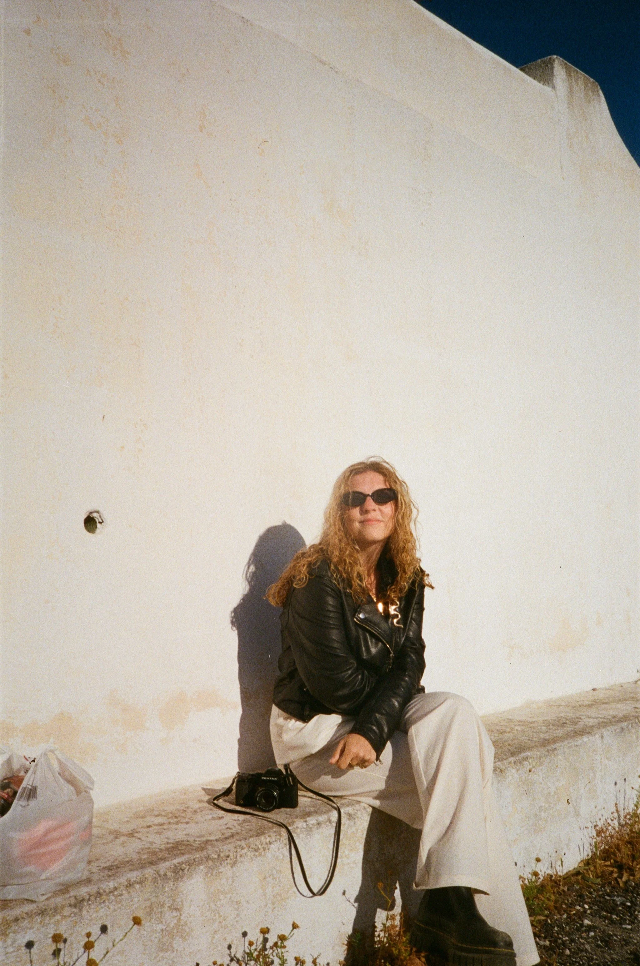 A woman with curly blonde hair, wearing black sunglasses, a black leather jacket, white pants, and black boots, sitting on a concrete ledge against a white wall with a small hole. She has a camera on the ledge beside her and is smiling. The scene is outdoors with some plants at the bottom.
