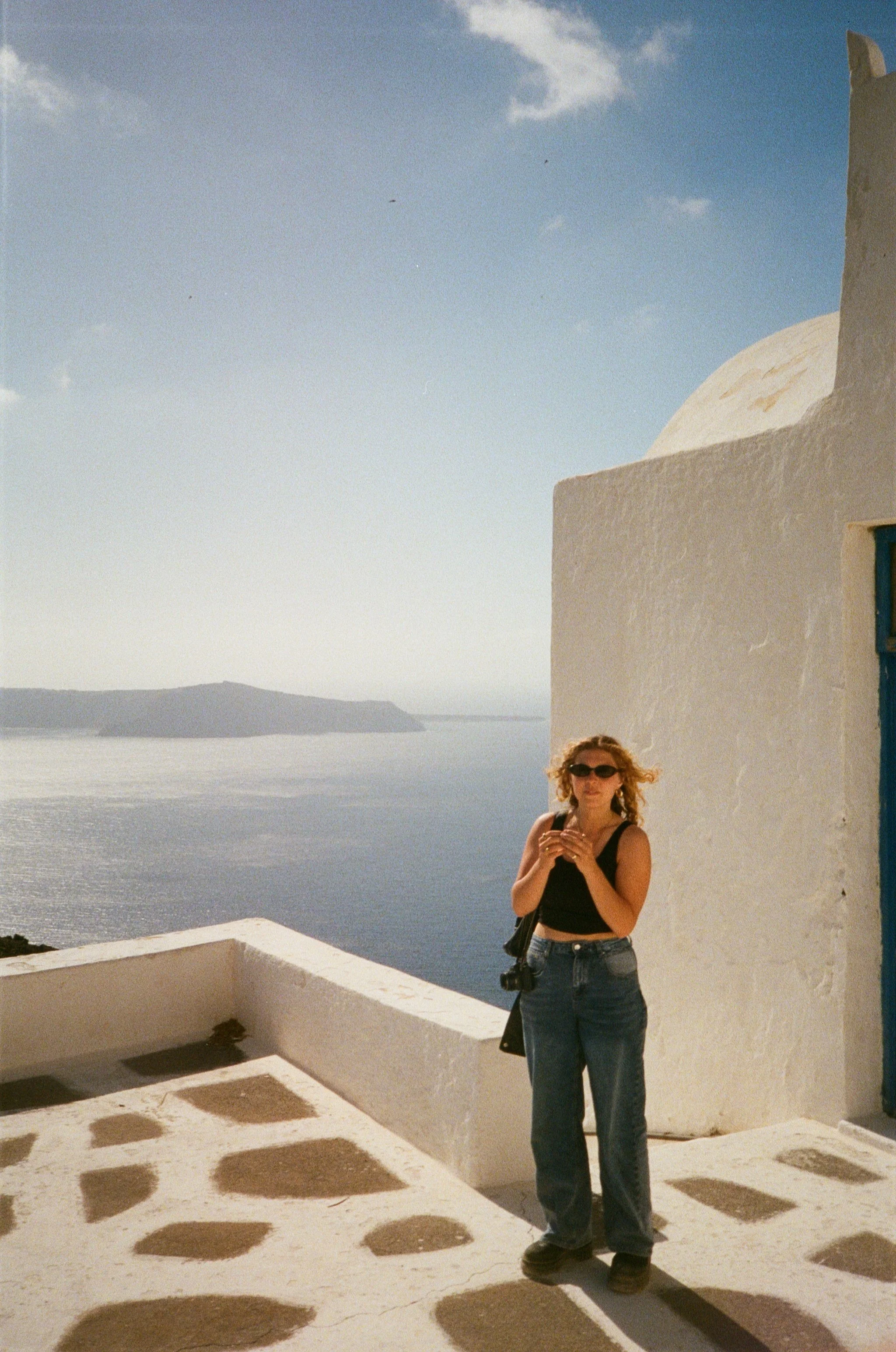 A woman with curly hair, wearing sunglasses, a black sleeveless top, and jeans, standing outside on a white balcony near a white building with a blue door, overlooking the ocean with distant land in the background, on a sunny day.