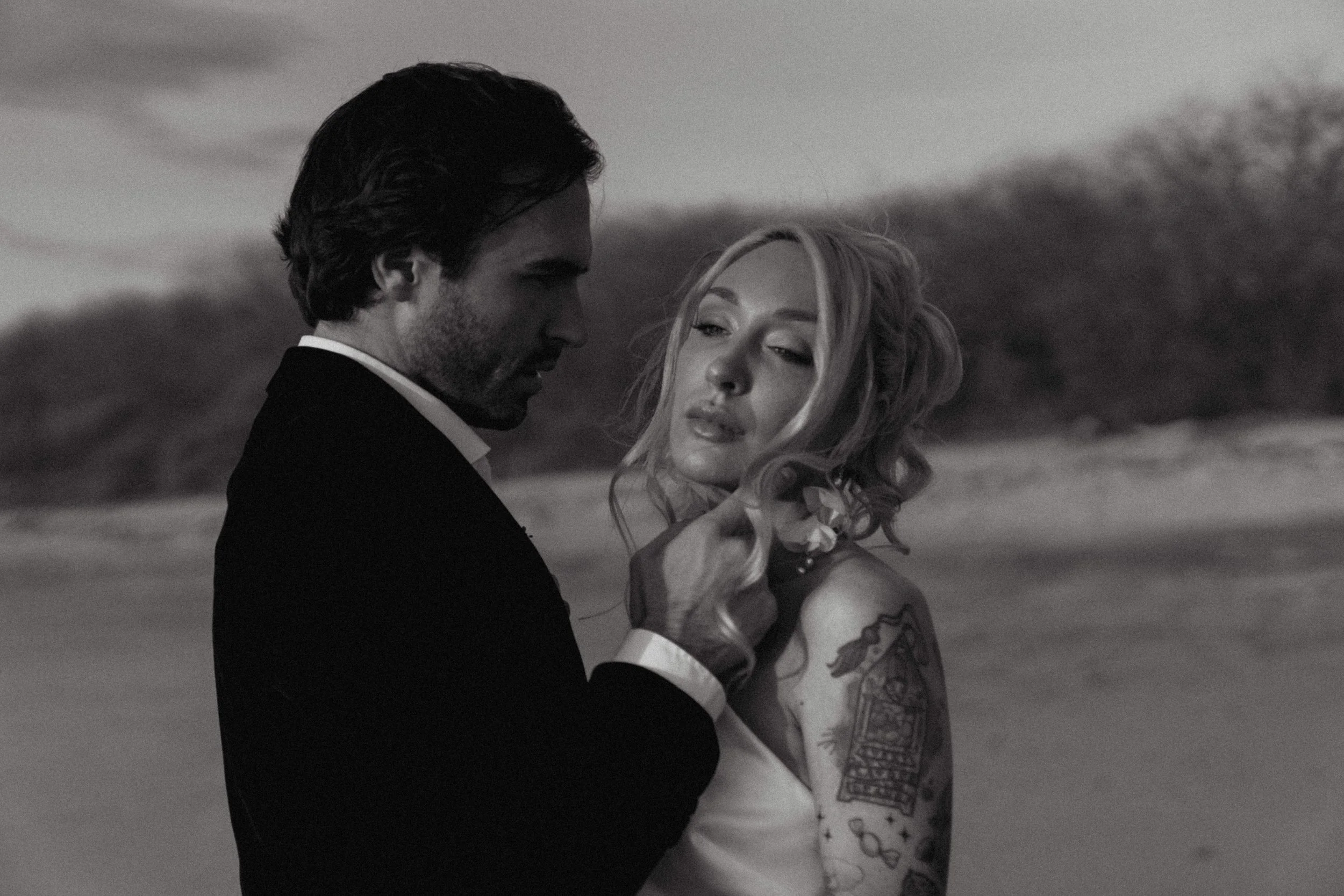 A black and white photo of a man and woman on a beach, with the man gently touching the woman's chin. The man has dark hair and a beard, wearing a suit, and the woman has blonde hair styled in loose curls, with tattoos on her arm, wearing a sleeveless dress.