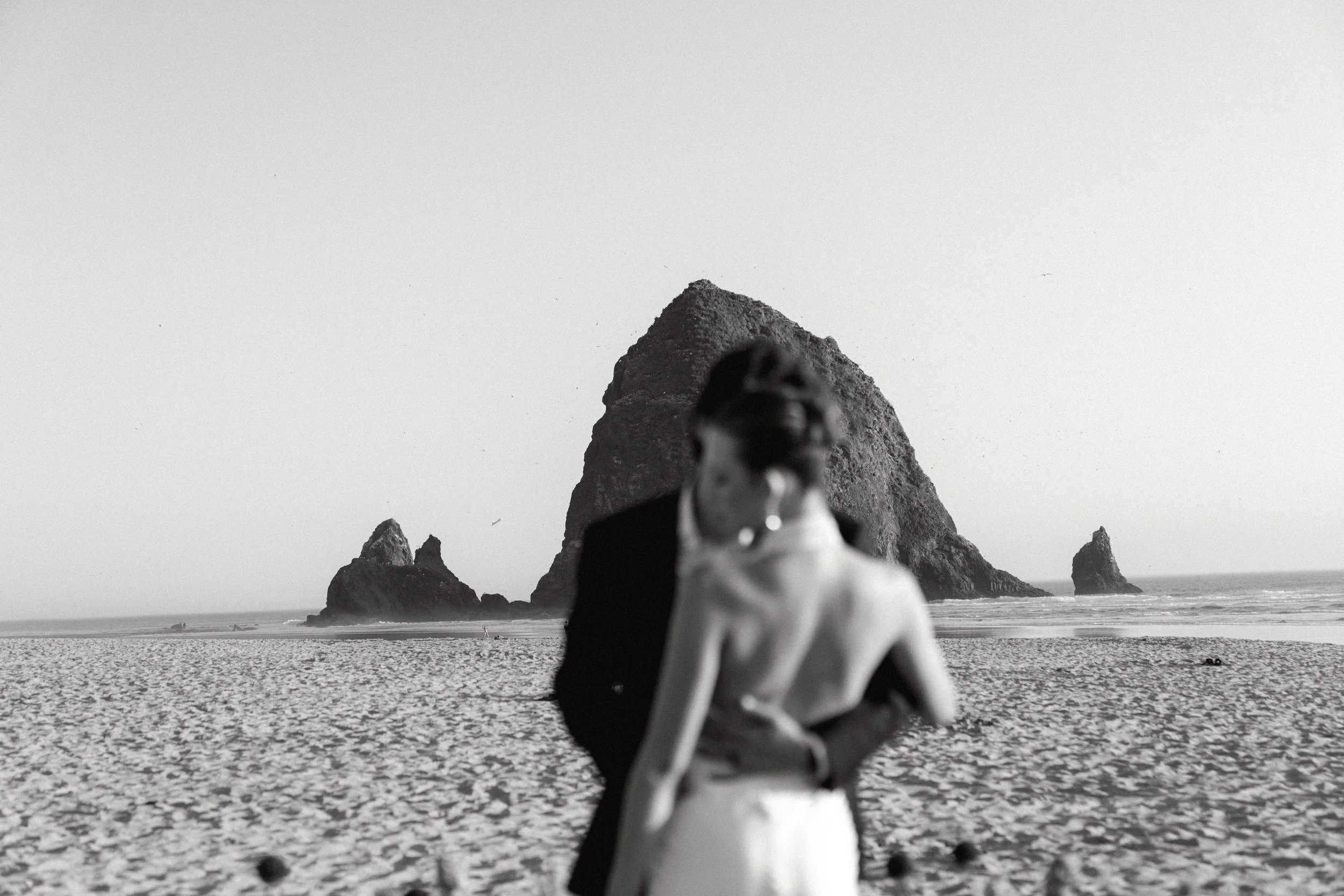 Bride and groom embrace on a beach at their elopement on the Oregon Coast. They are out of focus and in the background surrounding them is a large Haystack rock in the ocean with birds flying overhead. Black and White image.