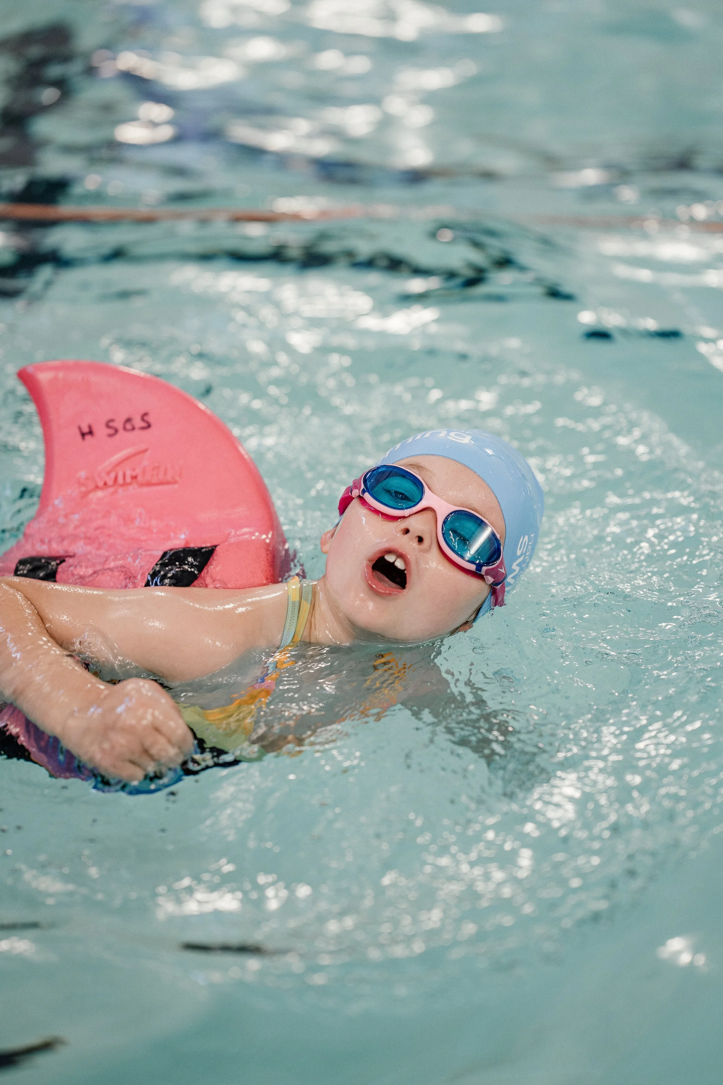 Young child wearing swimming goggles and a swim cap in a swimming pool, holding a pink kickboard, with water splashing around.