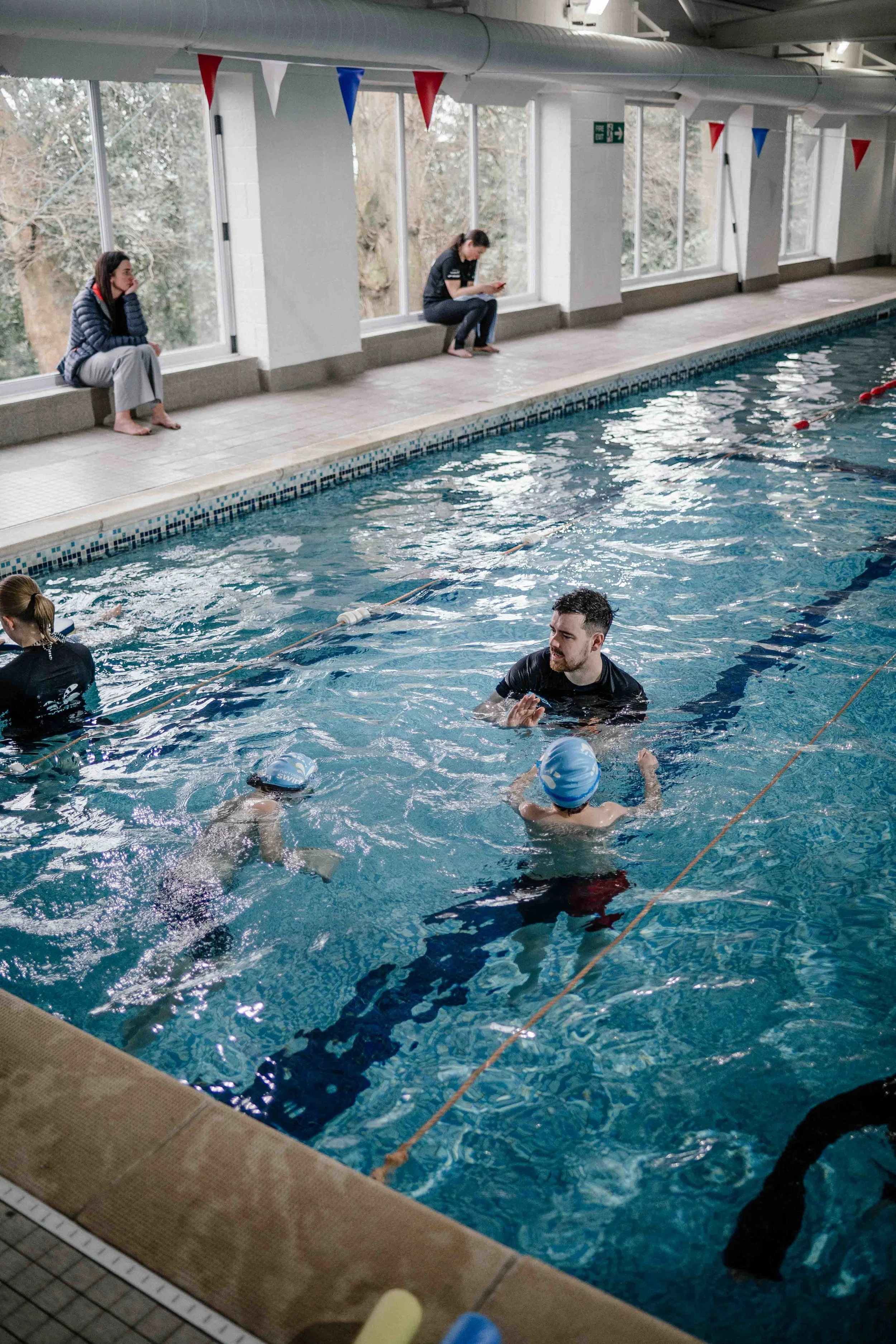 Indoor swimming pool with people swimming and sitting on the poolside, decorated with red, white, and blue bunting.