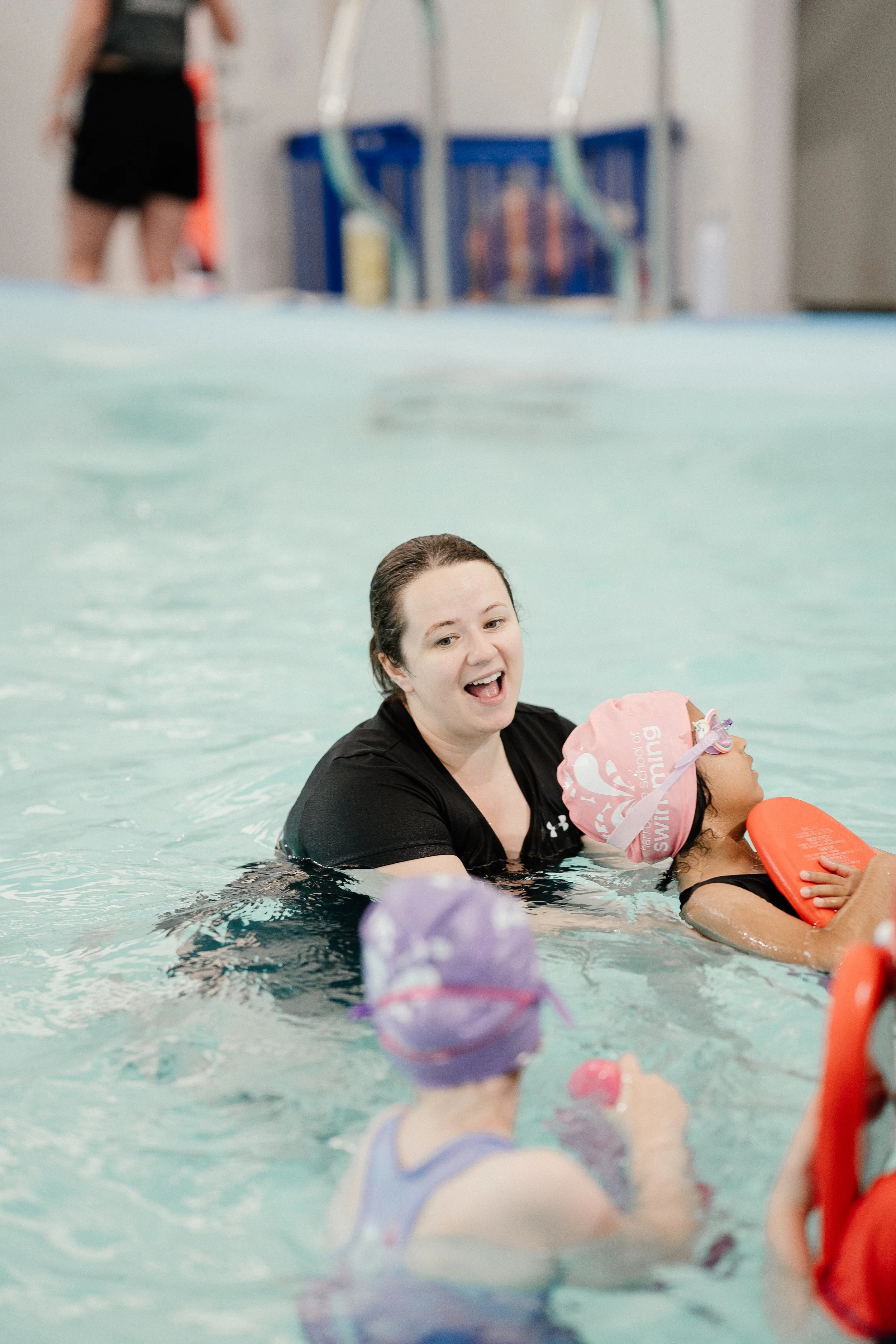 A woman assisting a young girl with swimming in a pool, both wearing swim caps and holding flotation devices.