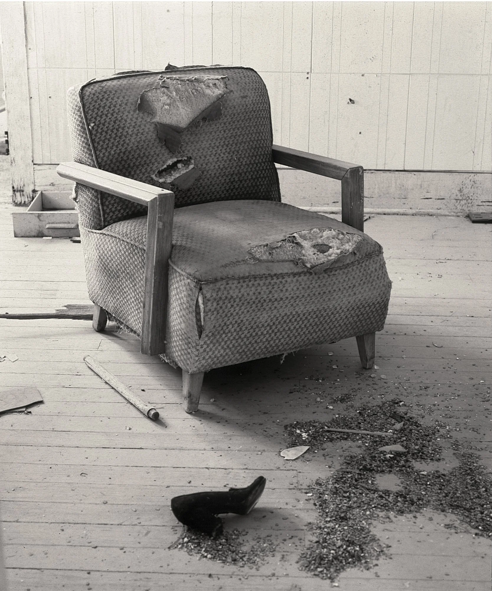 An old, worn-out armchair with missing fabric on the seat and backrest, exposing the cushioning inside. There is a wooden armrest on the left side, and the chair is placed on a wooden floor with dirt and debris scattered around. A broken black high-h