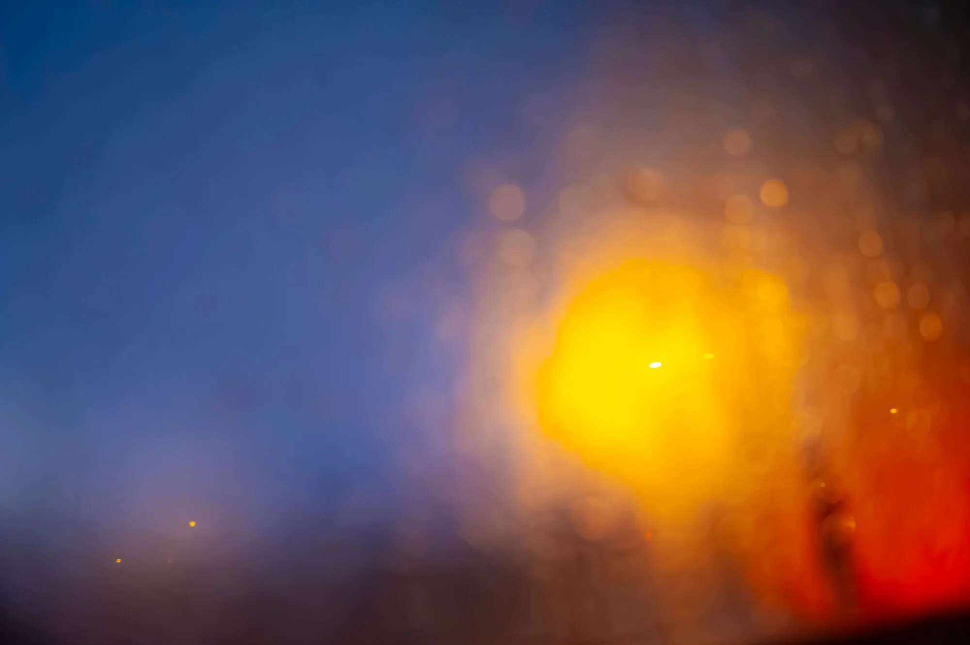 Blurred image of a bright yellow street light at night seen through rain-covered glass, creating bokeh effects with a dark blue and orange background.