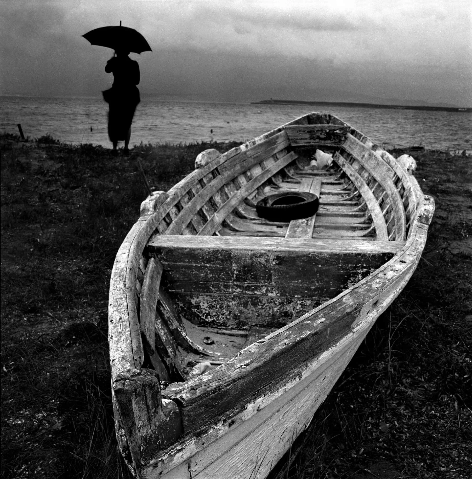 An old, weathered boat resting on the ground near a body of water, with a person holding an umbrella standing in the background on a cloudy day.