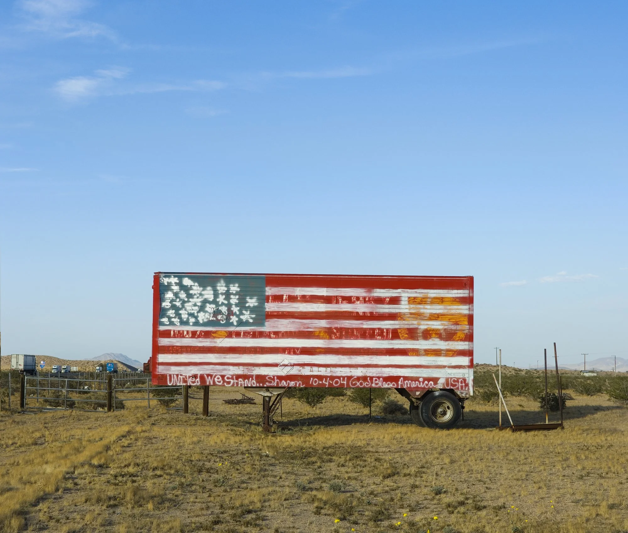 A trailer painted with a weathered American flag design in an open desert landscape under a blue sky with a few clouds.
