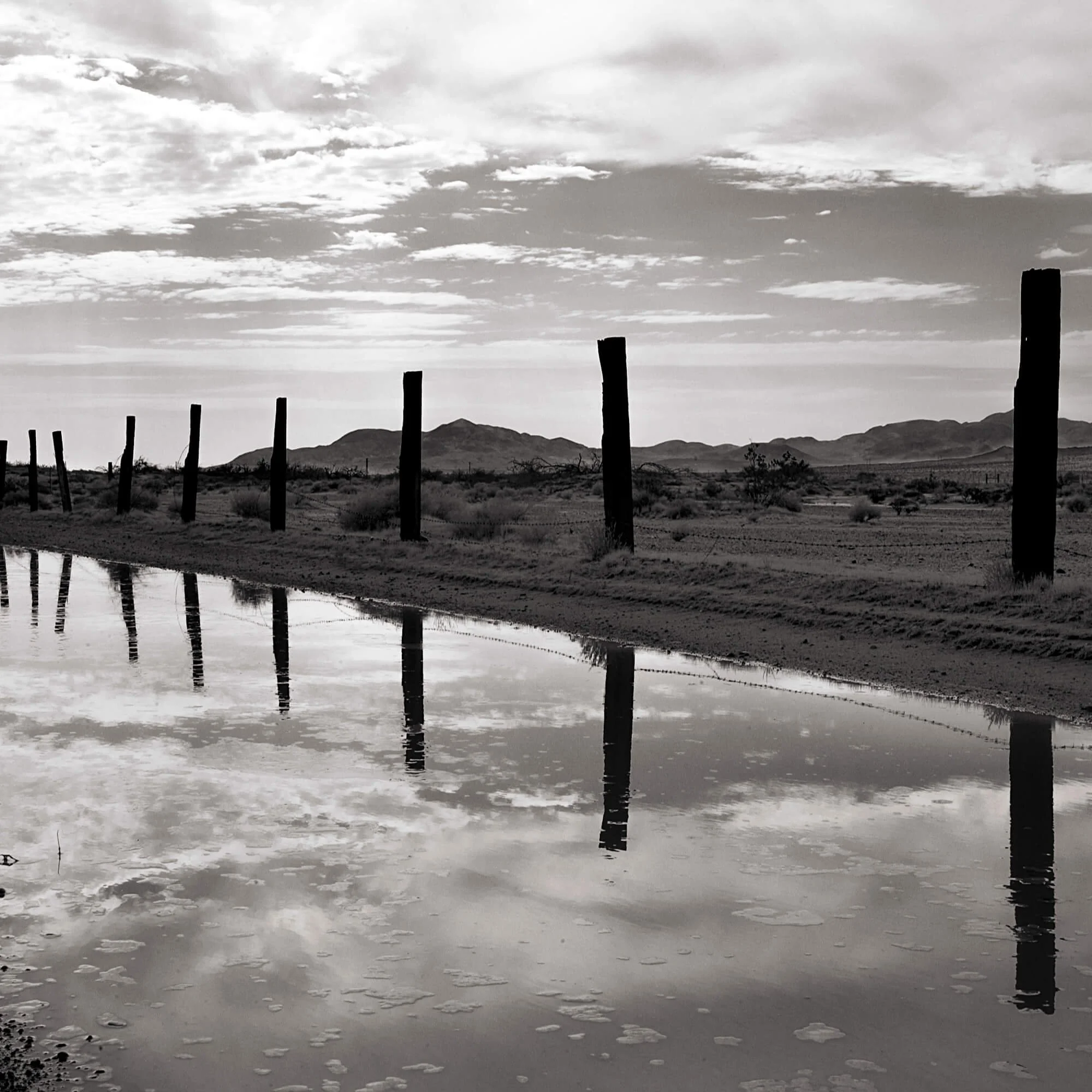 Black and white photograph of a desert landscape with mountain ranges in the background. Loose wooden fence posts run parallel along the horizon, and a body of water in the foreground reflects the fence posts and a partly cloudy sky.