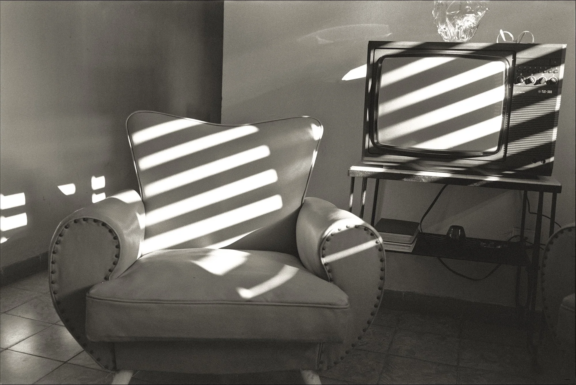 A vintage armchair with studded details is illuminated by sunlight passing through blinds, casting striped shadows. A small table with a vintage television and a glass bowl with decorative objects is in the background.