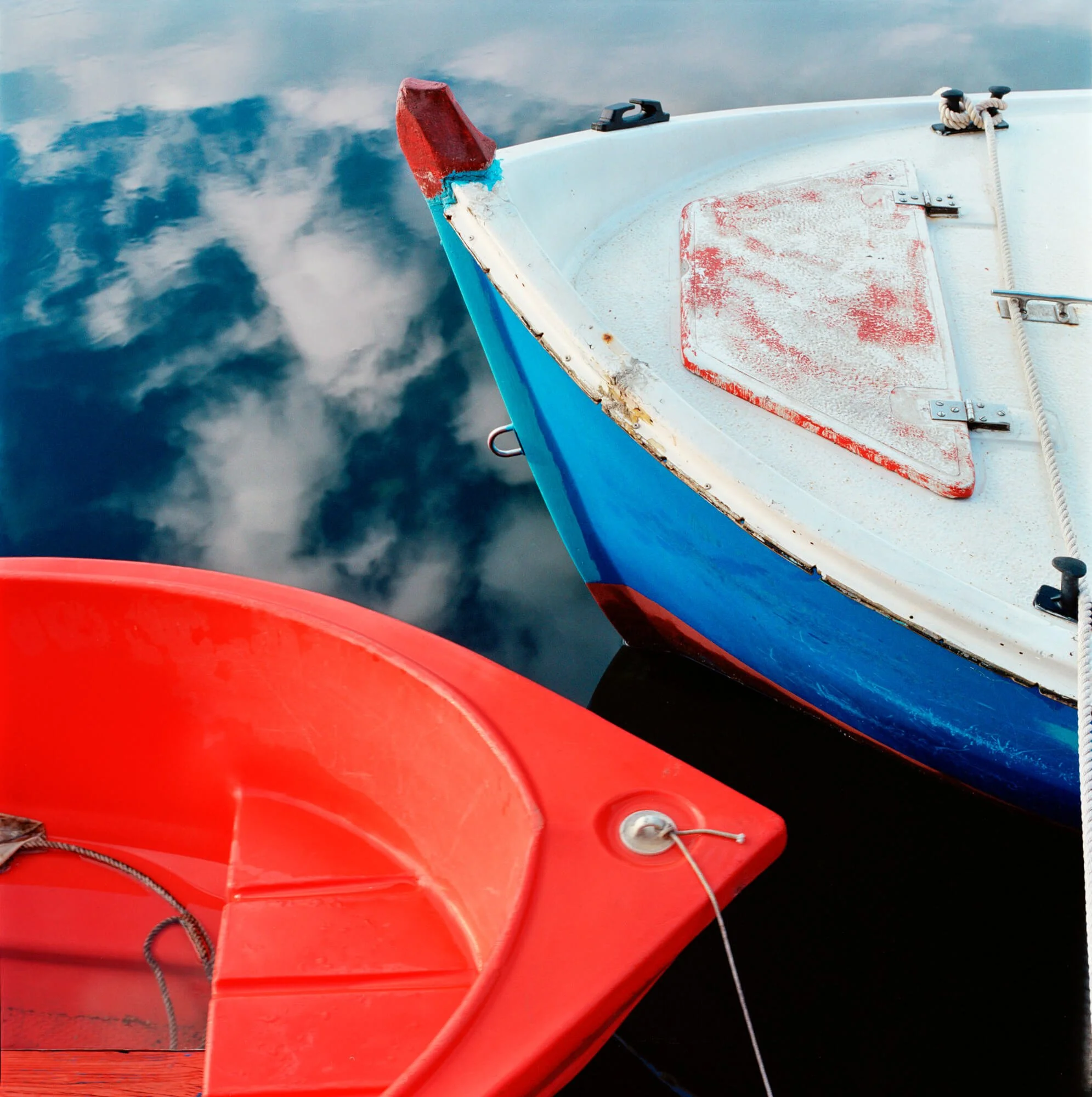 Close-up of two boats, one red and one blue, moored on calm water with reflections of clouds.