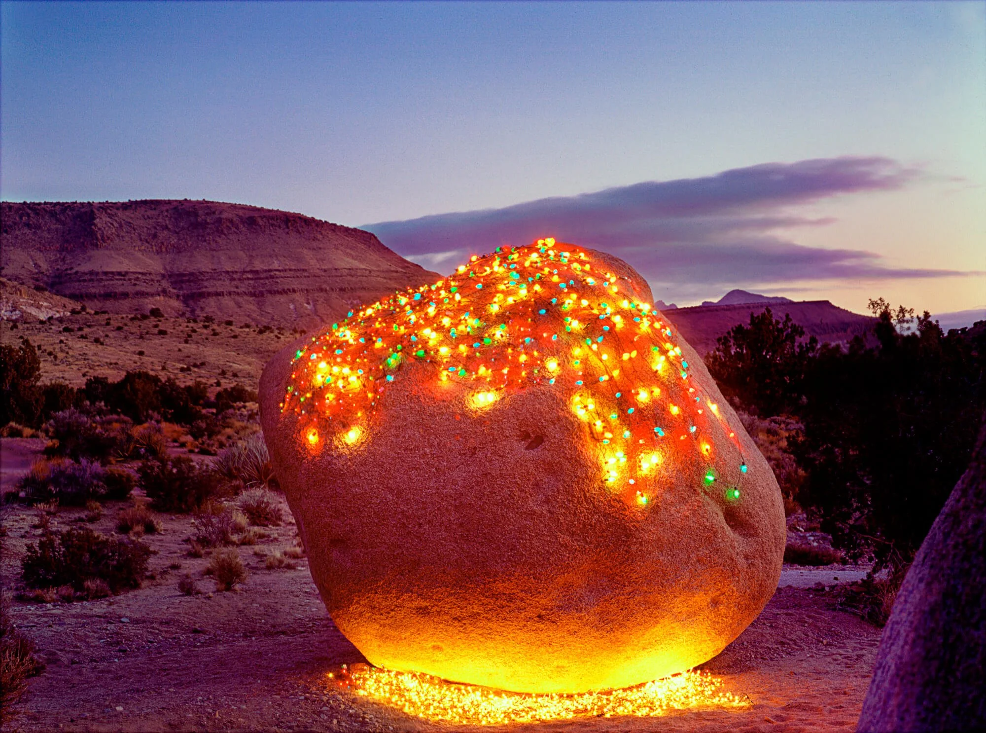 A large boulder with multicolored string lights on top, situated in a desert landscape at sunset with mountains and a cloudy sky in the background.