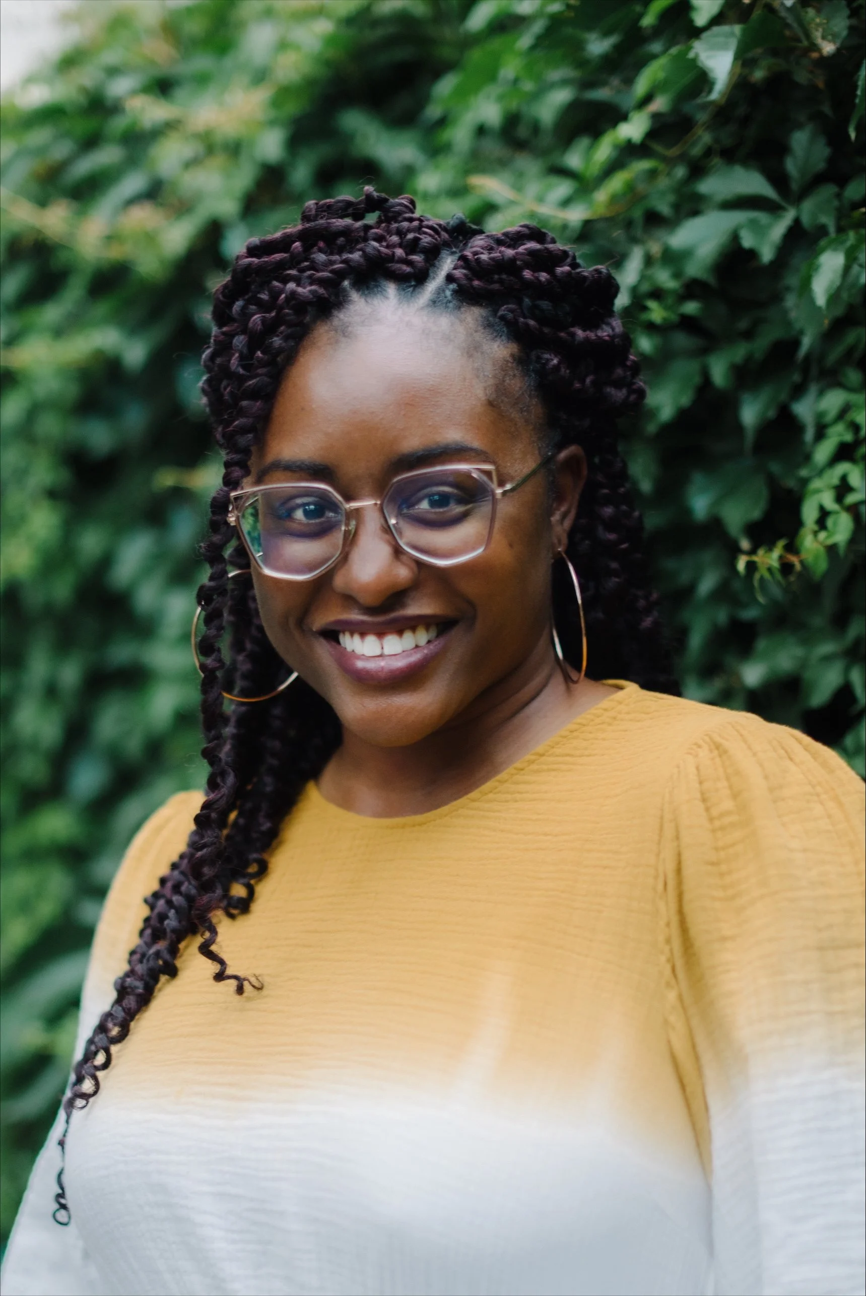 Smiling woman with glasses and hoop earrings, wearing a yellow and white top, standing outdoors in front of green foliage.