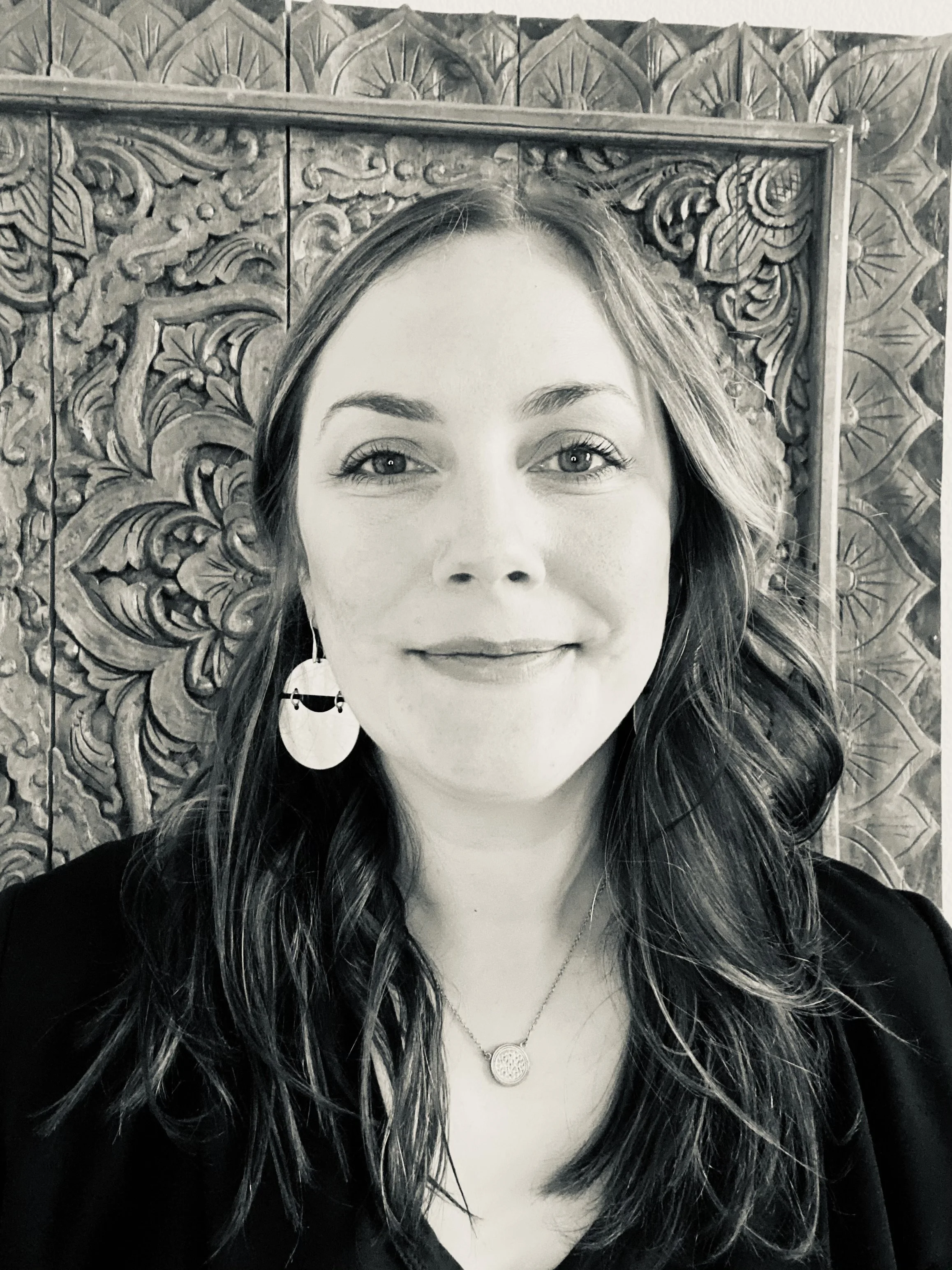 A woman smiling with long wavy hair, wearing earrings and a necklace, standing in front of an intricately carved wooden background.
