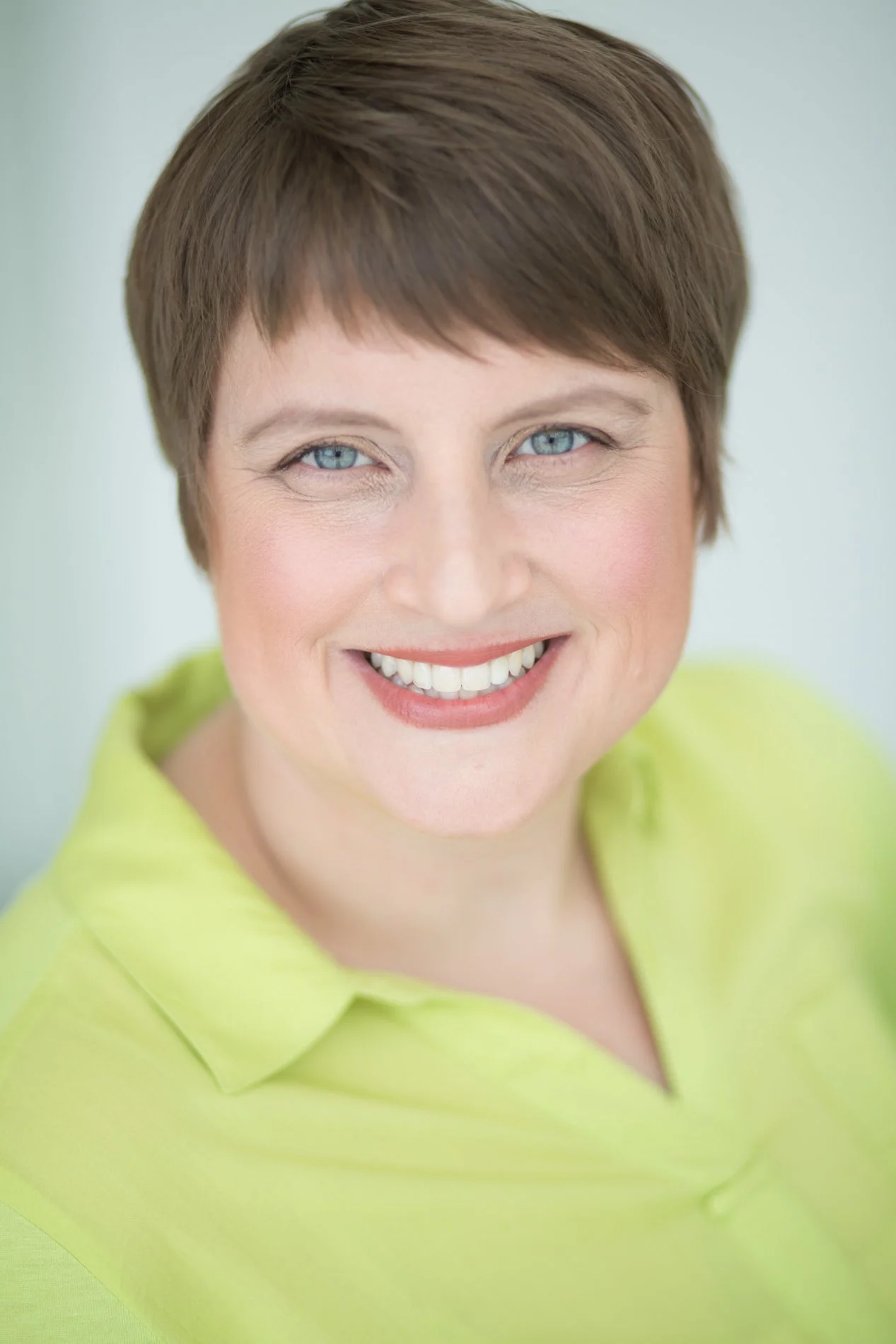 A woman with short brown hair and blue eyes smiling, wearing a bright yellow shirt.