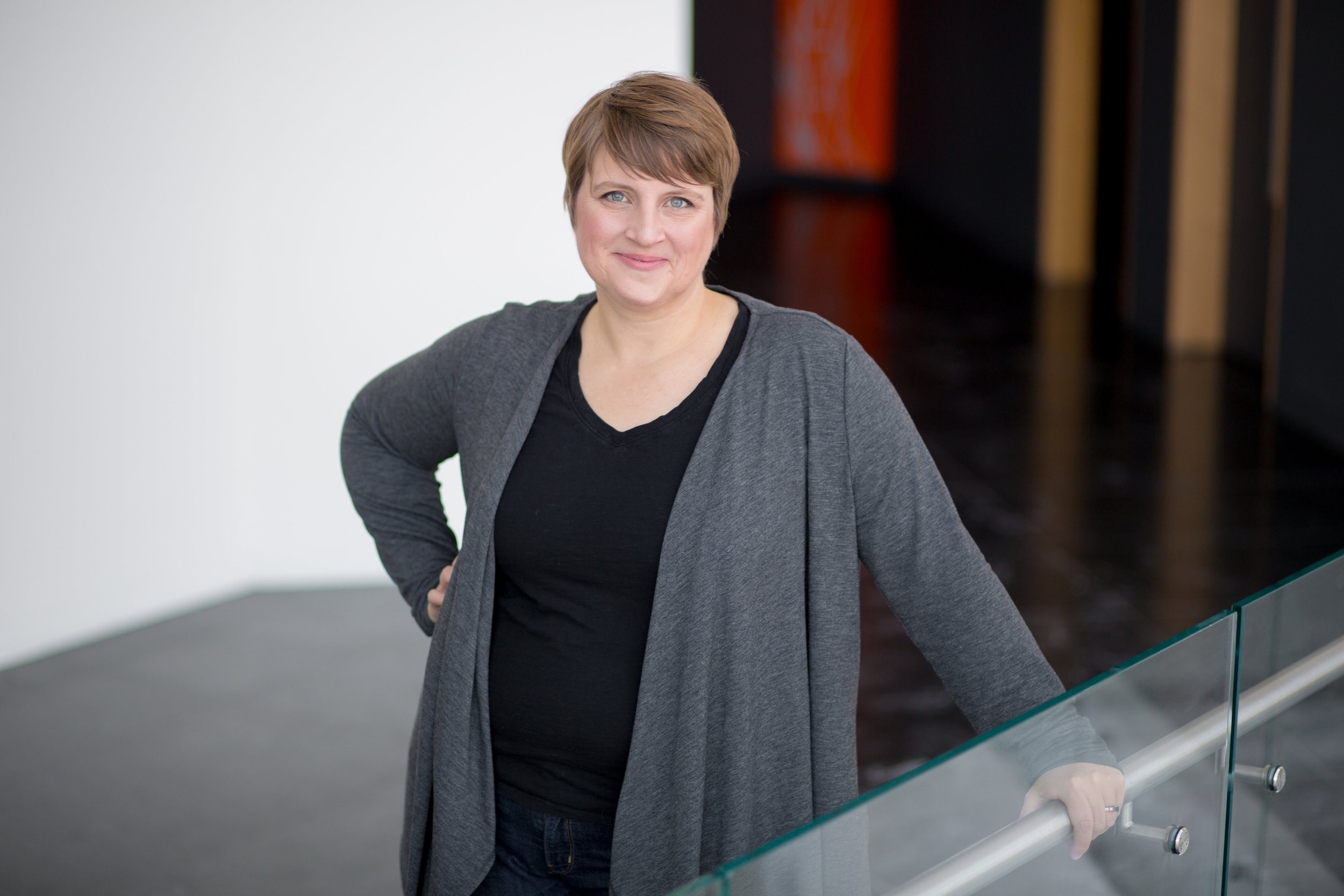 A woman with short brown hair and blue eyes wearing a gray blazer and black shirt, standing on a staircase with a glass railing in a modern building.