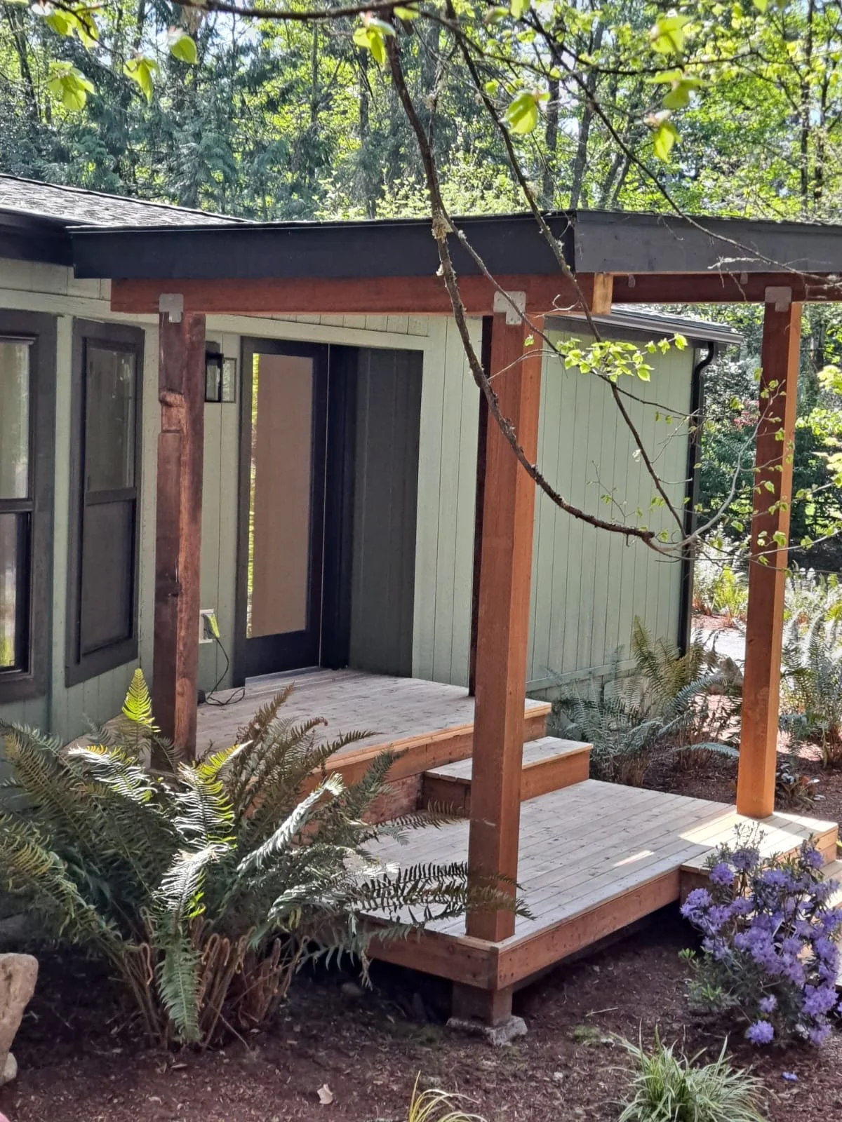 A small wooden porch attached to a house with steps leading up to a door, surrounded by lush greenery and purple flowers.