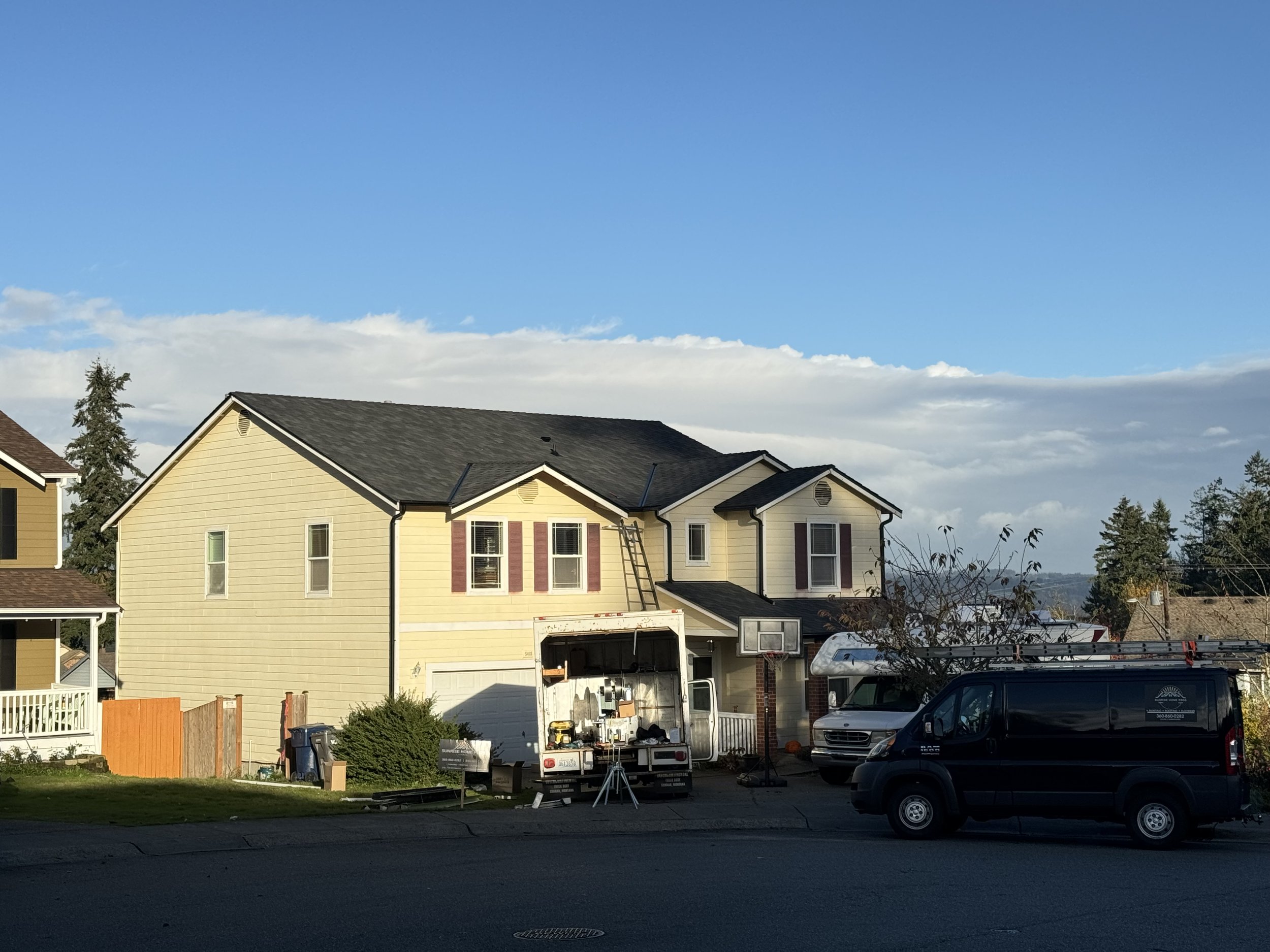 Residential house with maintenance vehicle and ladder, basketball hoop, and parked vans, clear blue sky with clouds.
