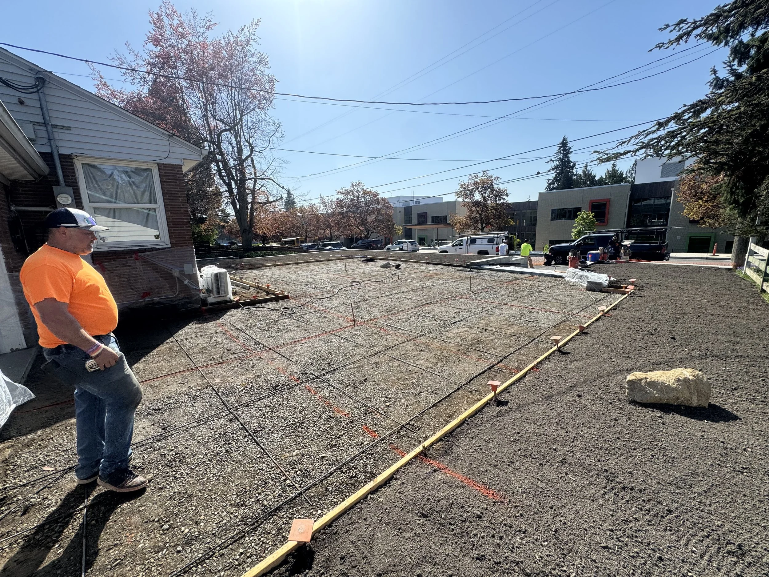 Construction site with a person in an orange shirt observing the foundation framework, and others working in the background on a sunny day.