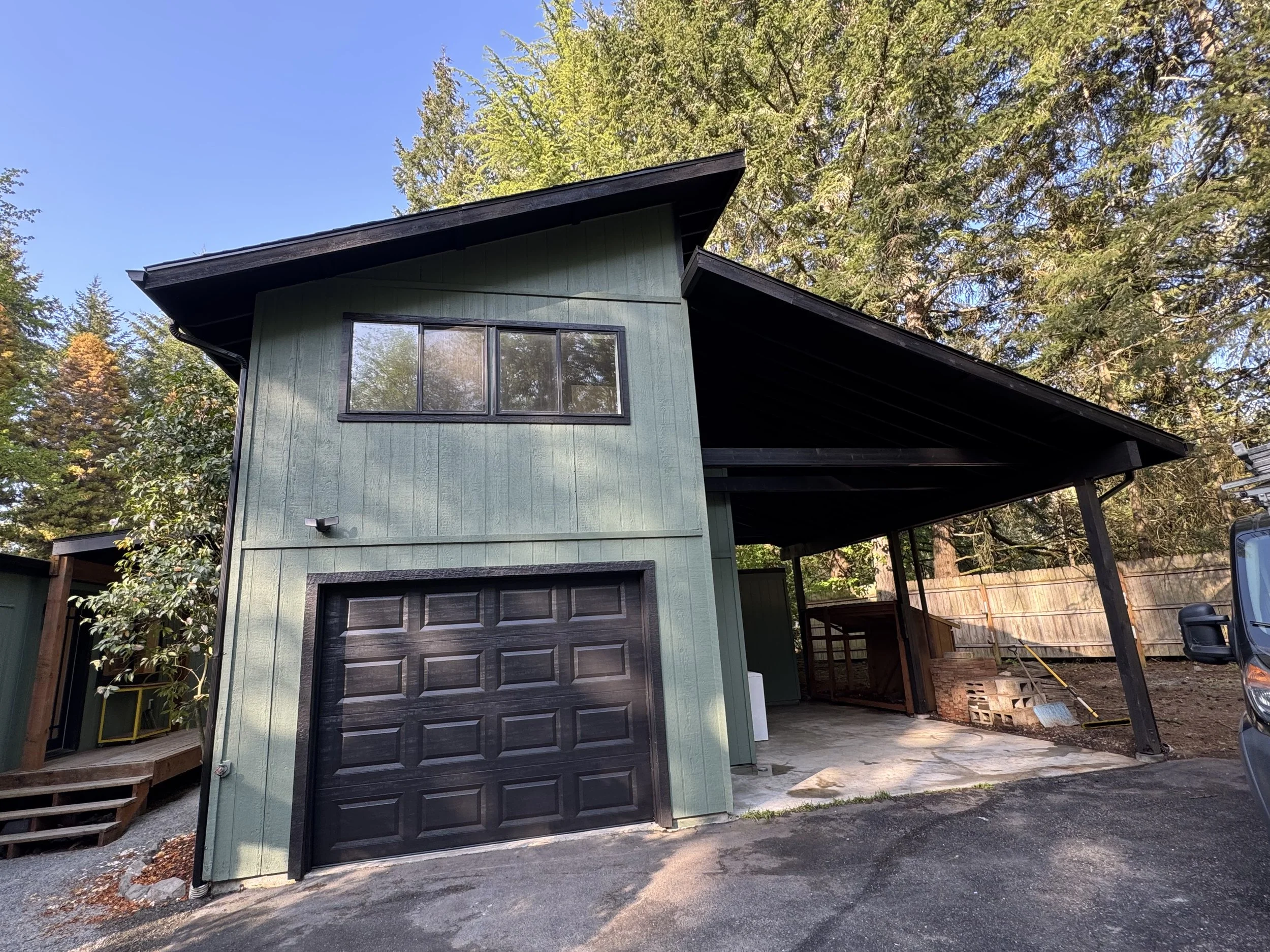 Modern green house with black garage door and large window, surrounded by trees, with a wooden fence and the driveway in the foreground.