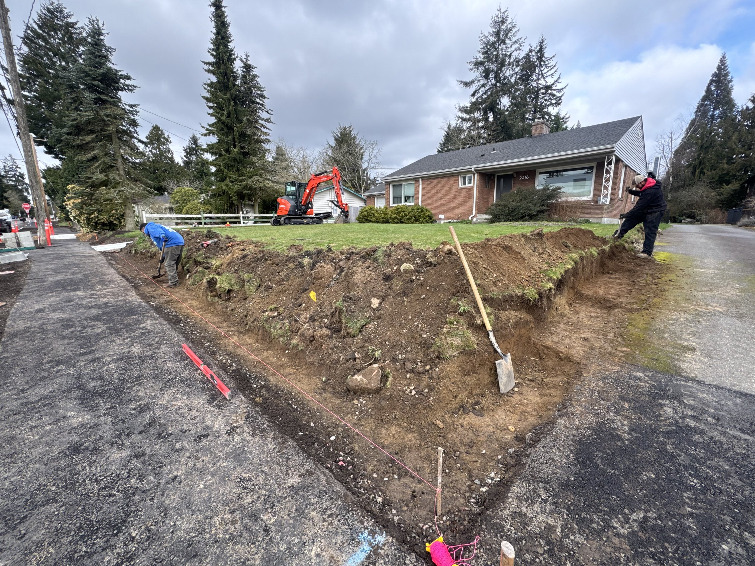 Construction workers digging a trench near a residential house, with a mini excavator parked on the grass, and the house in the background.