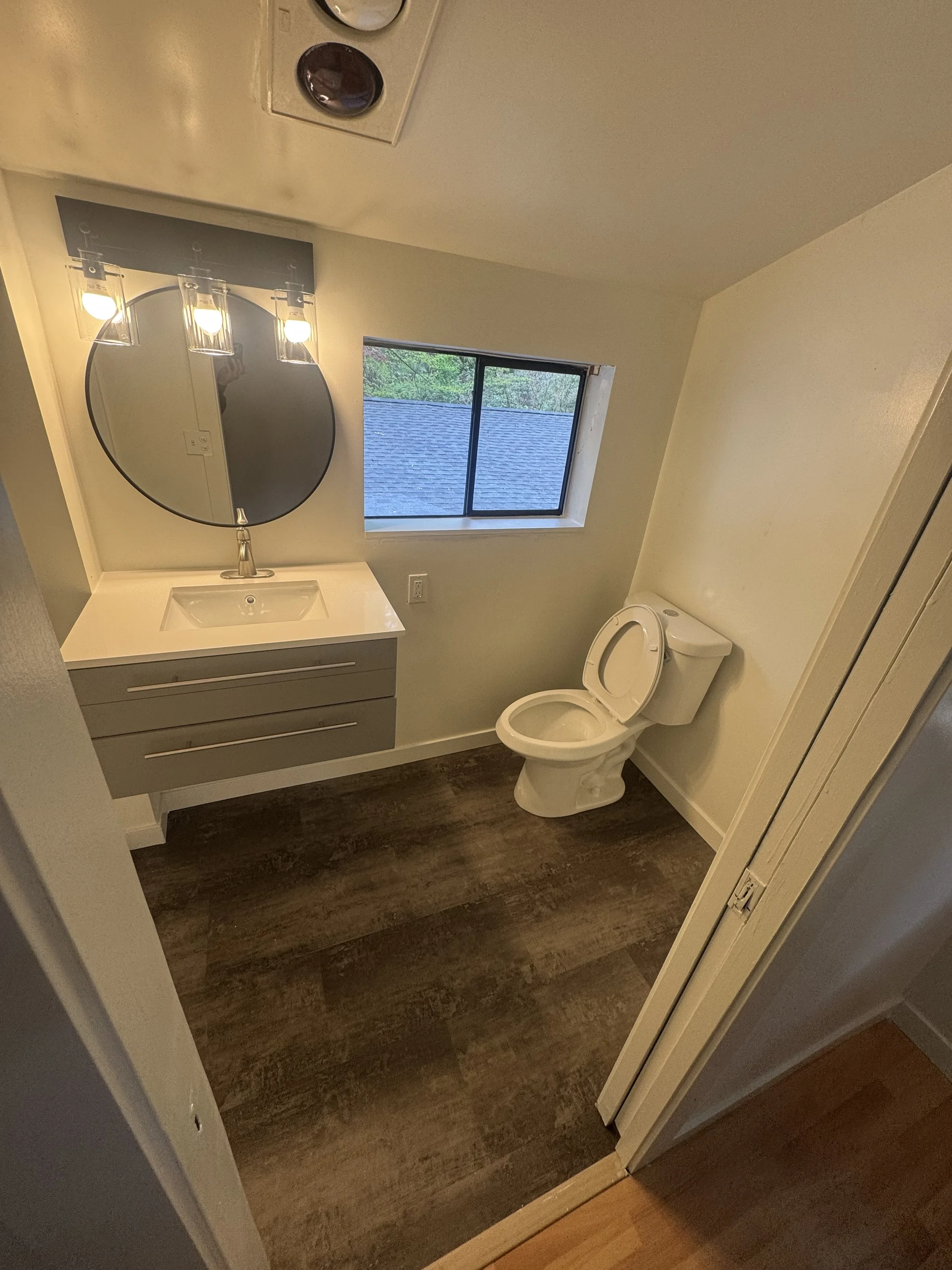 Small bathroom with a white toilet and a floating vanity with a round mirror, a window showing a roof and trees outside, and wood flooring.