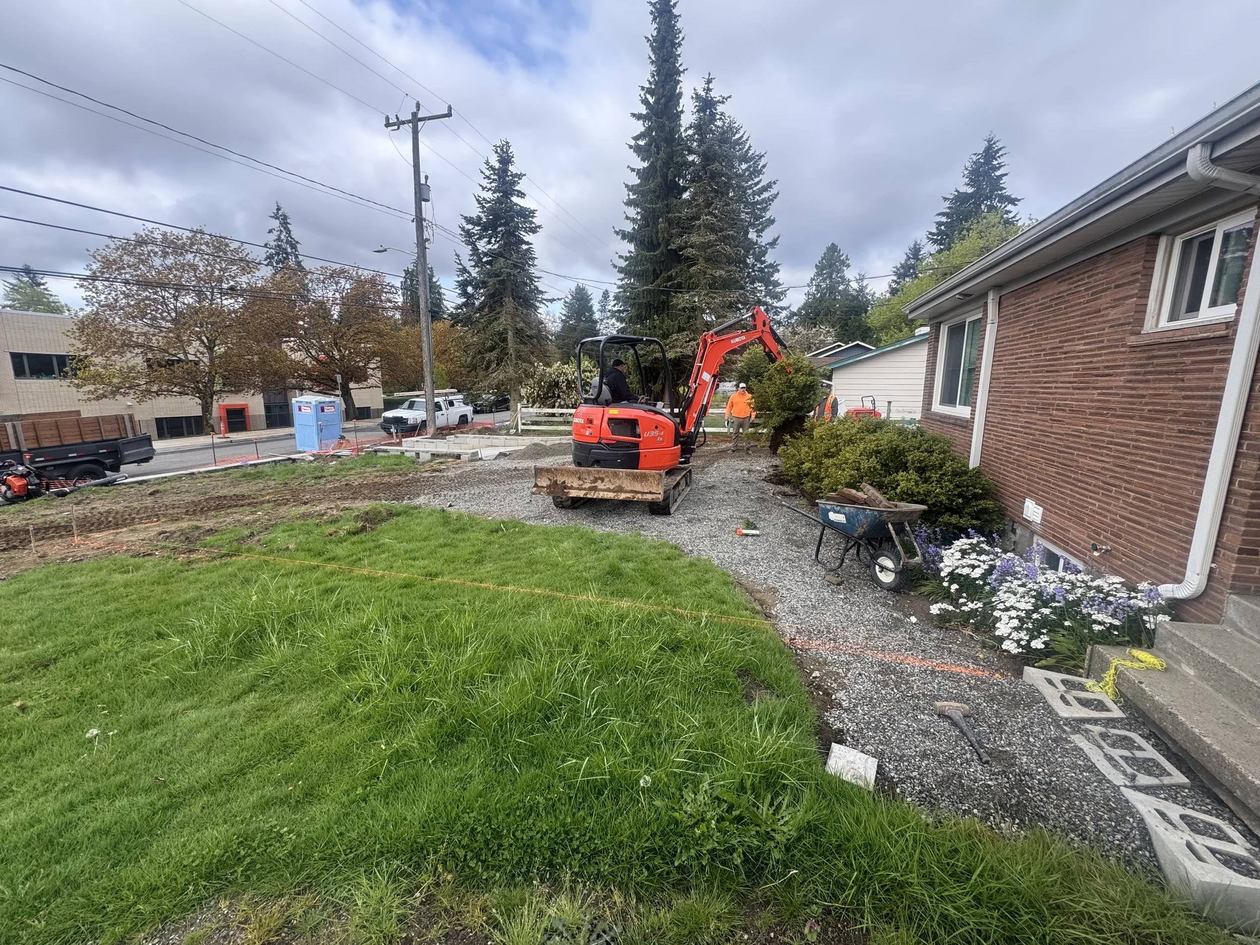 Construction site in a residential yard with a small red excavator, workers, flower plants, and building materials.