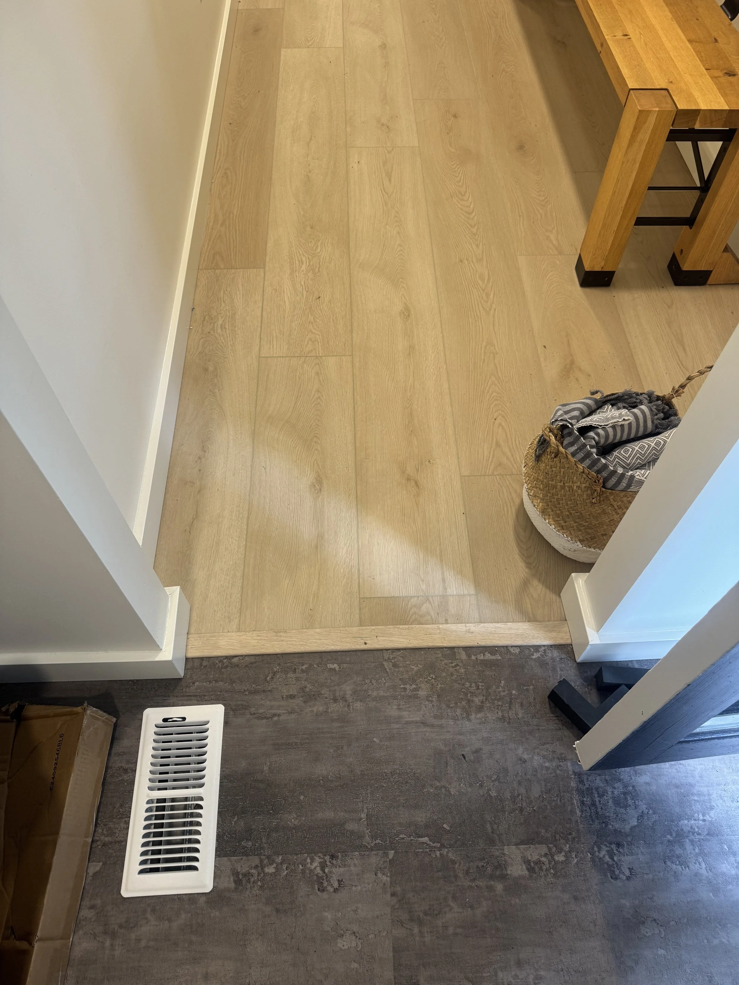 Transition between dark gray and light wood flooring in a home interior, with a white air vent on the gray floor and a wicker basket with black and gray cloths on the wood floor near a wooden table.