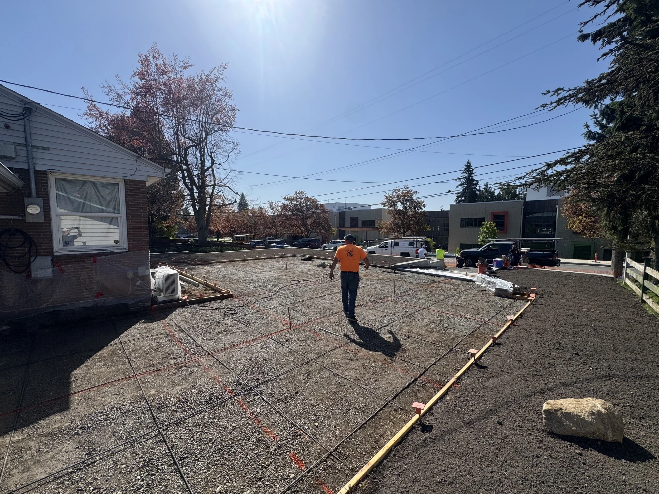 Construction site with a worker walking on a gravel foundation, marked with red and yellow outline, in front of a building and trees with autumn foliage under a clear blue sky.