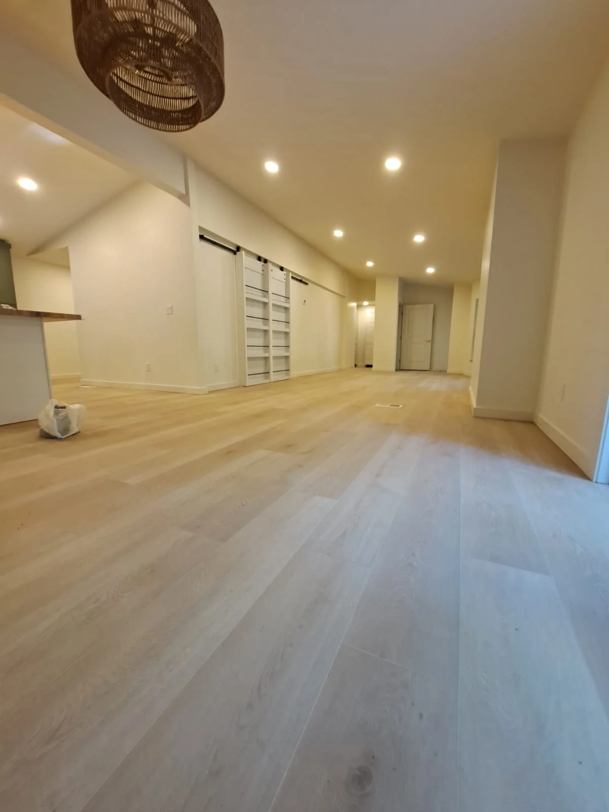 Empty living room with light-colored wooden flooring, white walls, white sliding barn door, ceiling lights, living room area, and a hanging wicker light fixture.