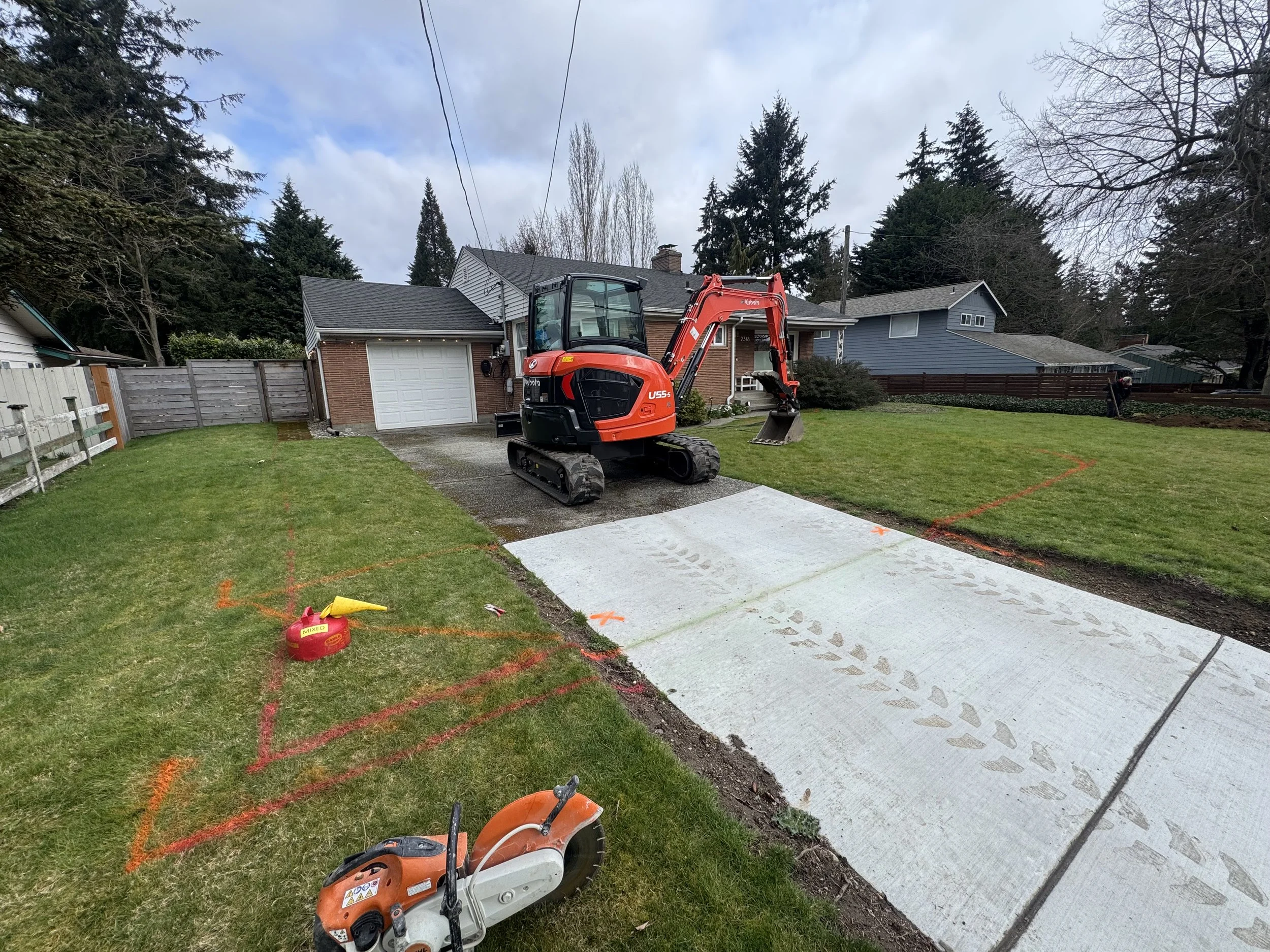Residential backyard with new concrete patio being installed; construction equipment including a mini excavator, a small saw, and marking spray in the yard; orange markings and markings on the concrete indicate construction layout.
