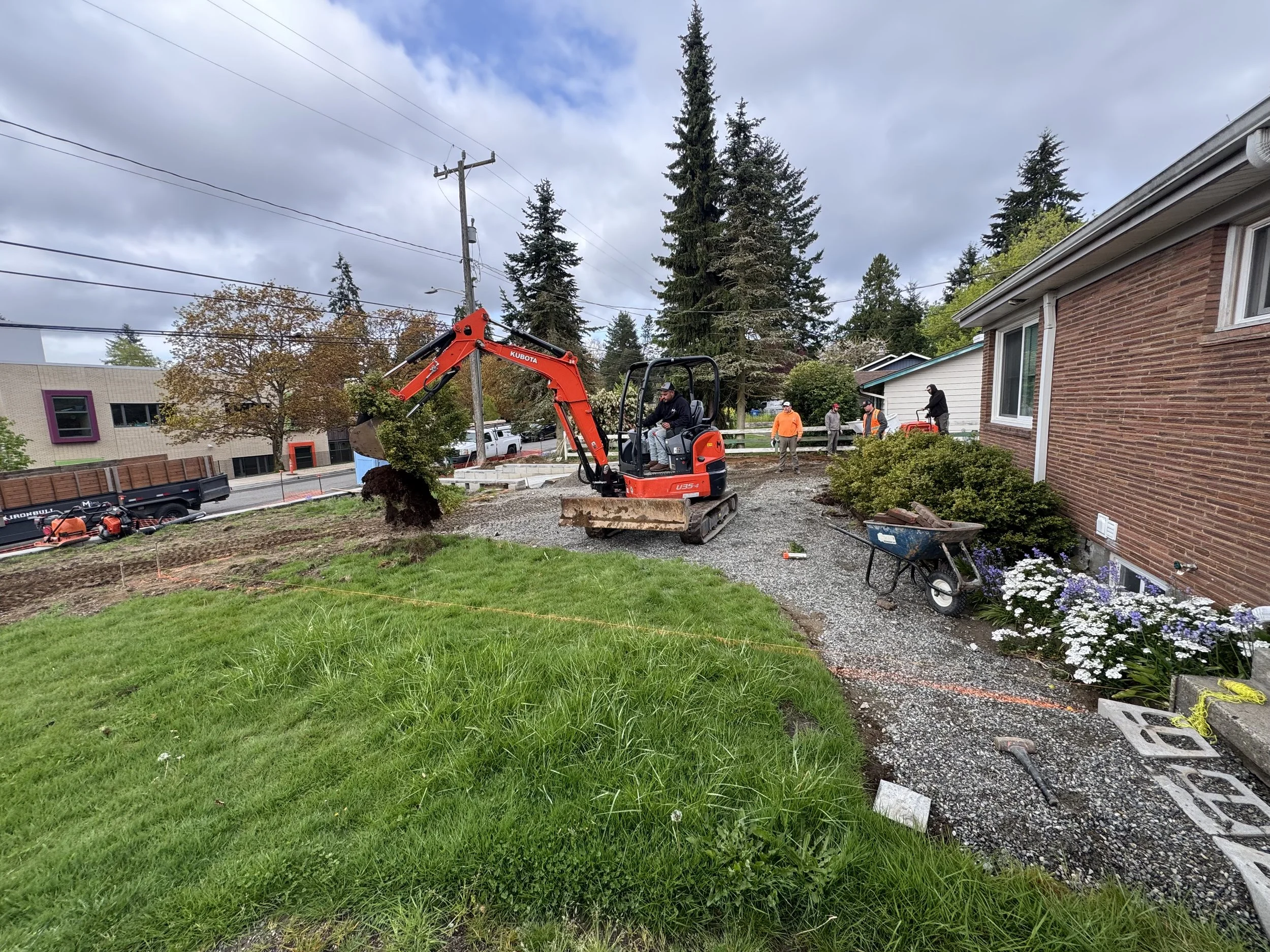 Construction workers and a small excavator digging up soil near a house with a brick exterior, with trees, flowers, and power lines in the background.