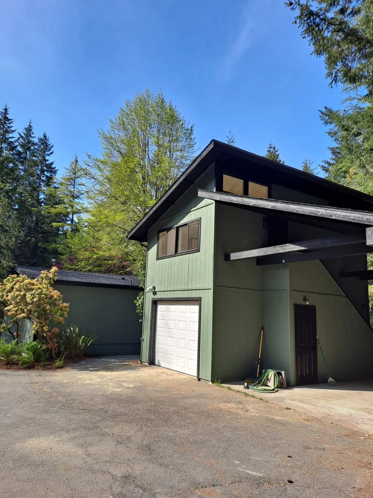 A green two-story house with a white garage door, dark brown door, and black staircase on the side, surrounded by trees and a bright blue sky.
