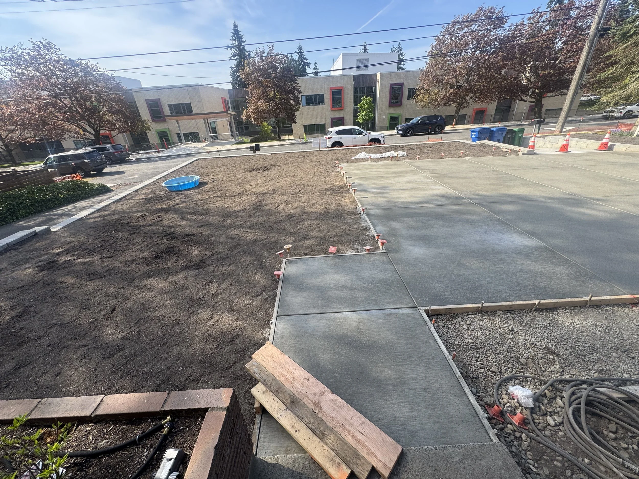 Construction site with freshly laid concrete sidewalk, dirt area, and construction cones, with parking lot and modern building in the background.