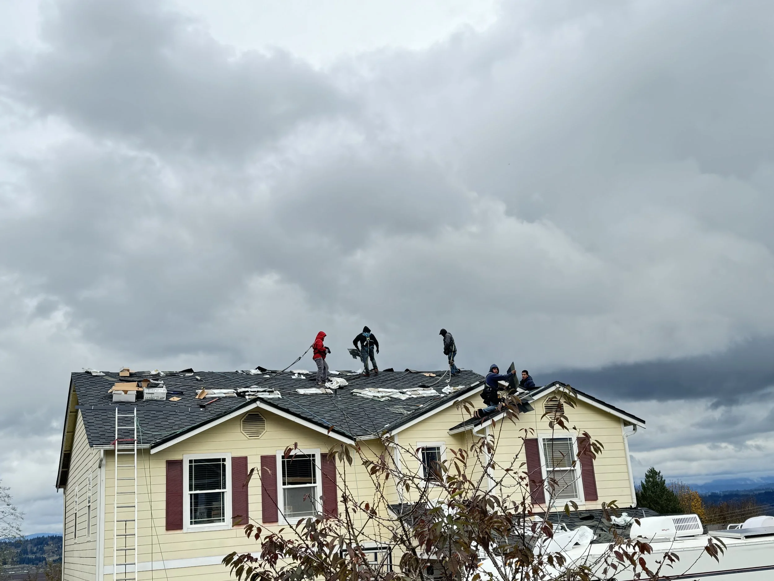 People working on a house roof in a cloudy sky, some kneeling and others standing, with tools and materials scattered around.