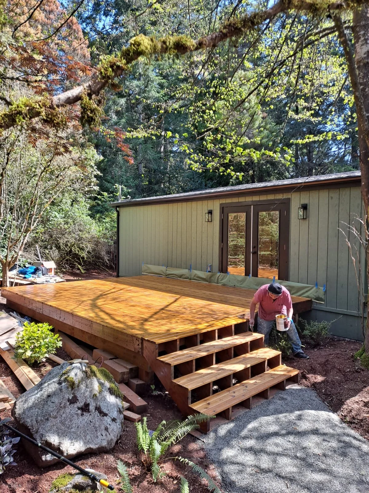 A person working on installing a wooden deck in a backyard, with a house and trees in the background.