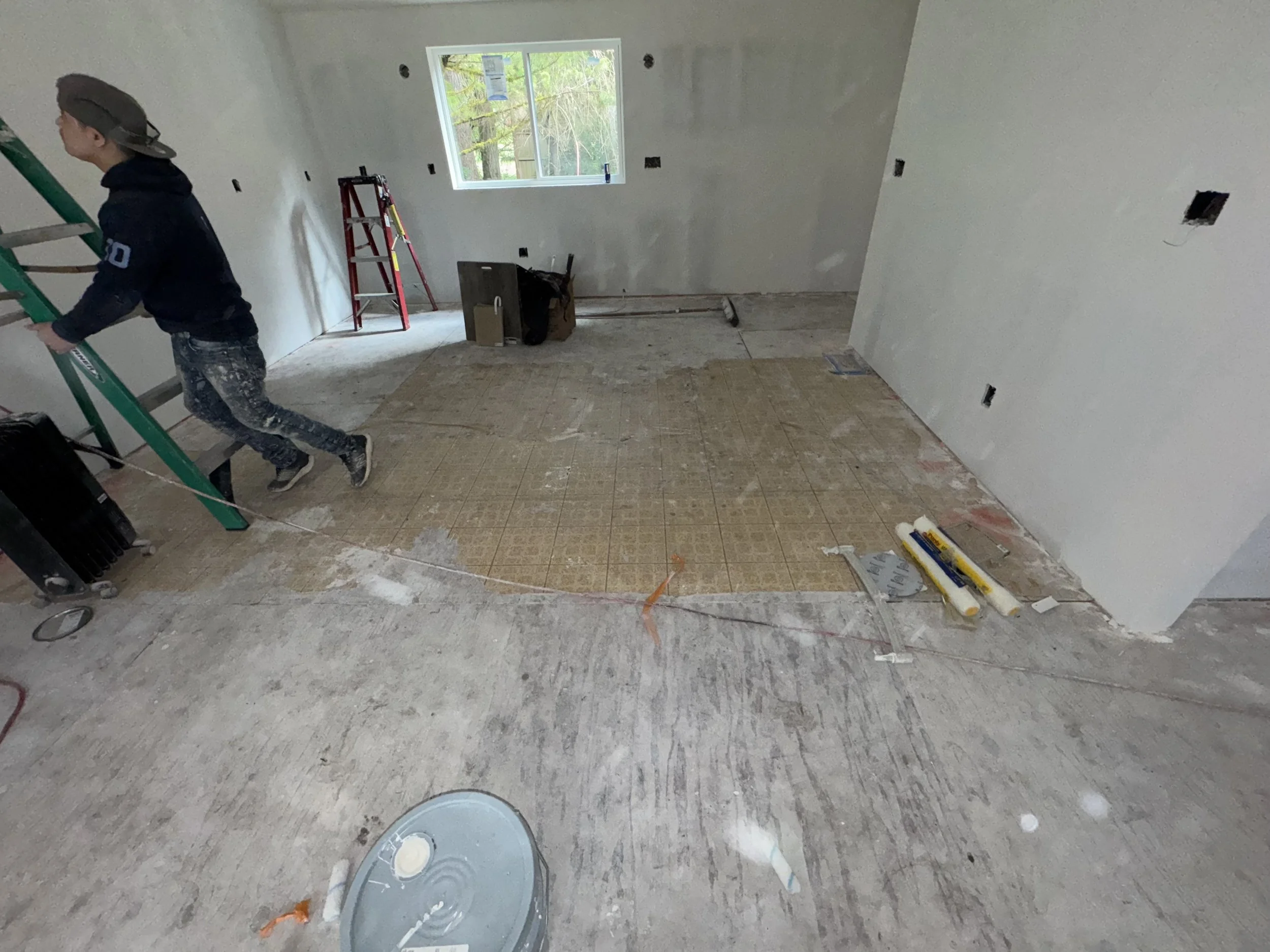 A person in construction clothing working in a room under renovation, with unfinished walls, a window, and construction tools and materials on the floor.