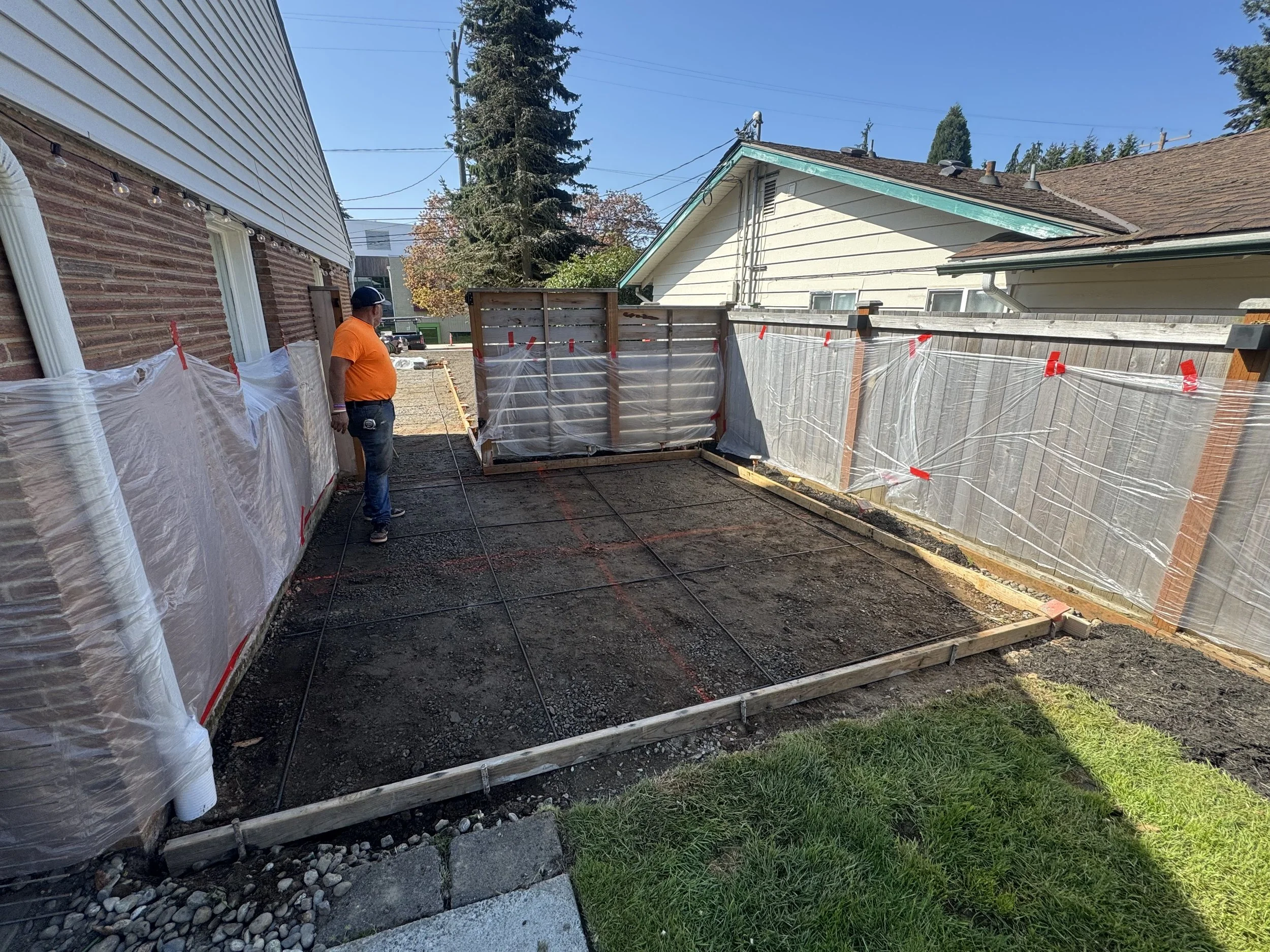 A man in an orange shirt and blue jeans stands in a backyard with a partially constructed patio, with a wooden frame and grid lines marked on the ground. The yard is enclosed by a wooden fence, and neighboring houses are visible in the background.