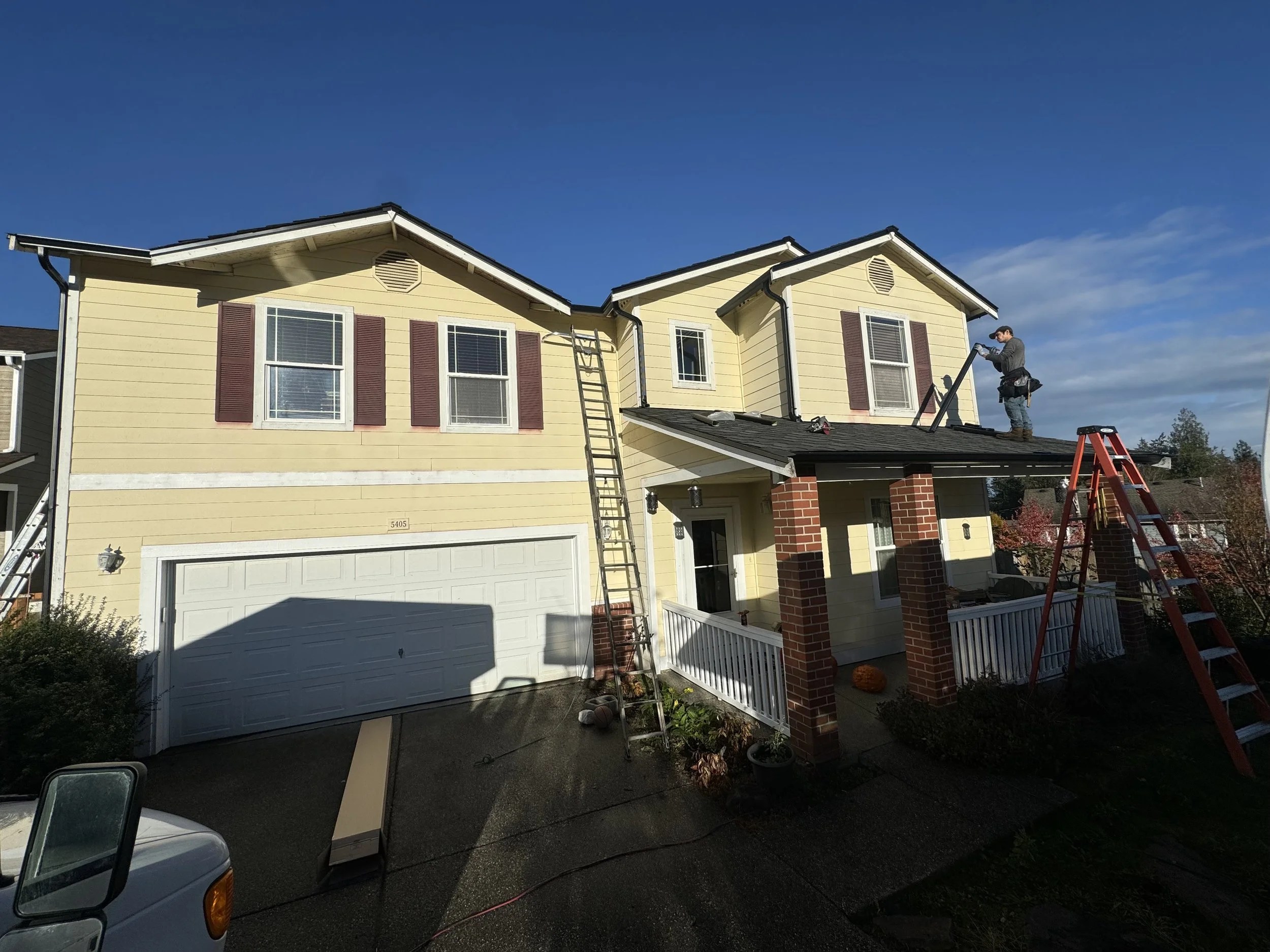 A two-story yellow house with white trim and red shutters undergoing roof repairs. Two workers are on the roof, with ladders leaning against the house, and a bright blue sky overhead.