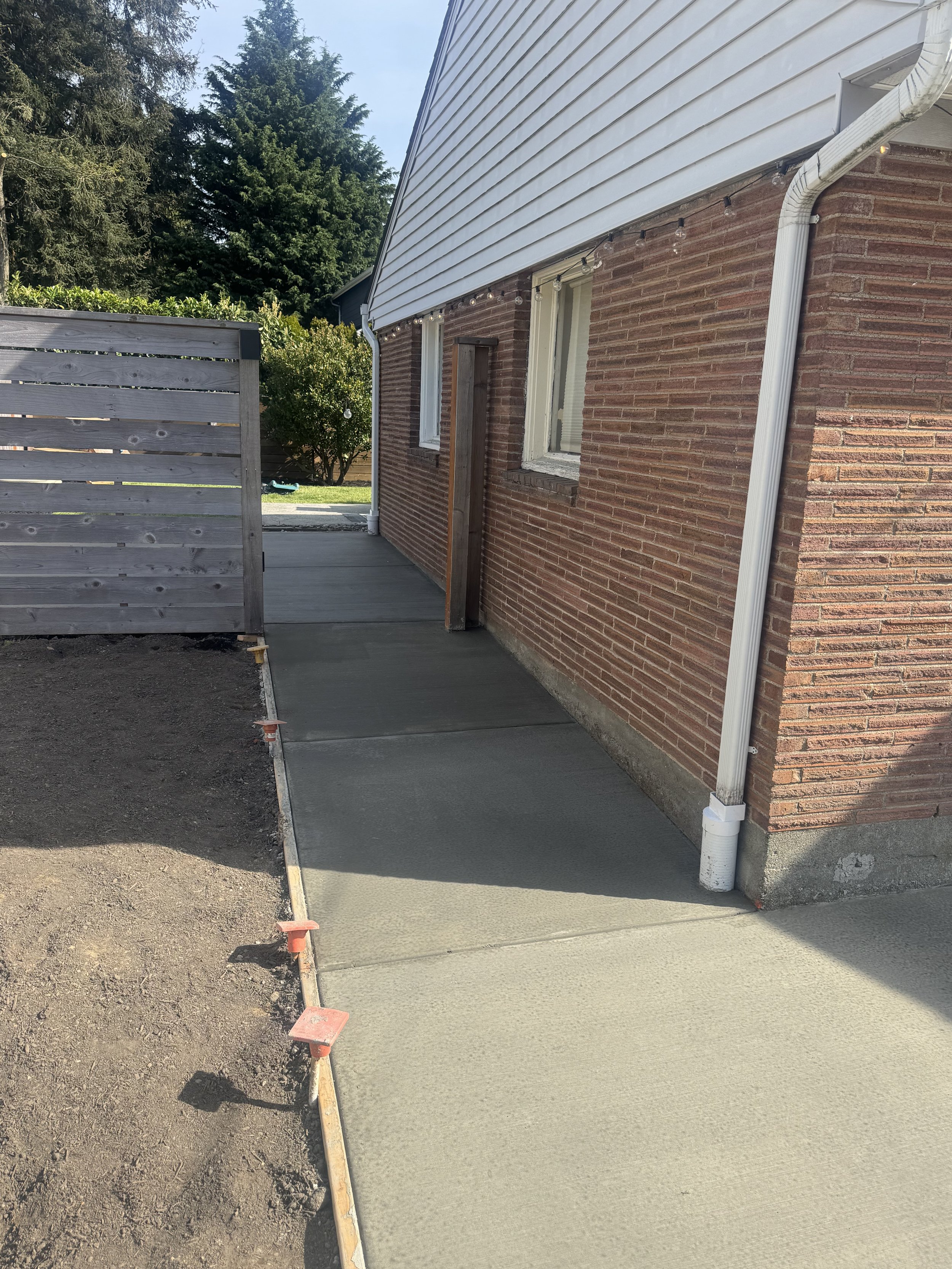 Concrete sidewalk being poured alongside a brick house with white siding on the upper part, white gutter, and two windows. There is a wooden privacy fence on the left, and trees in the background.