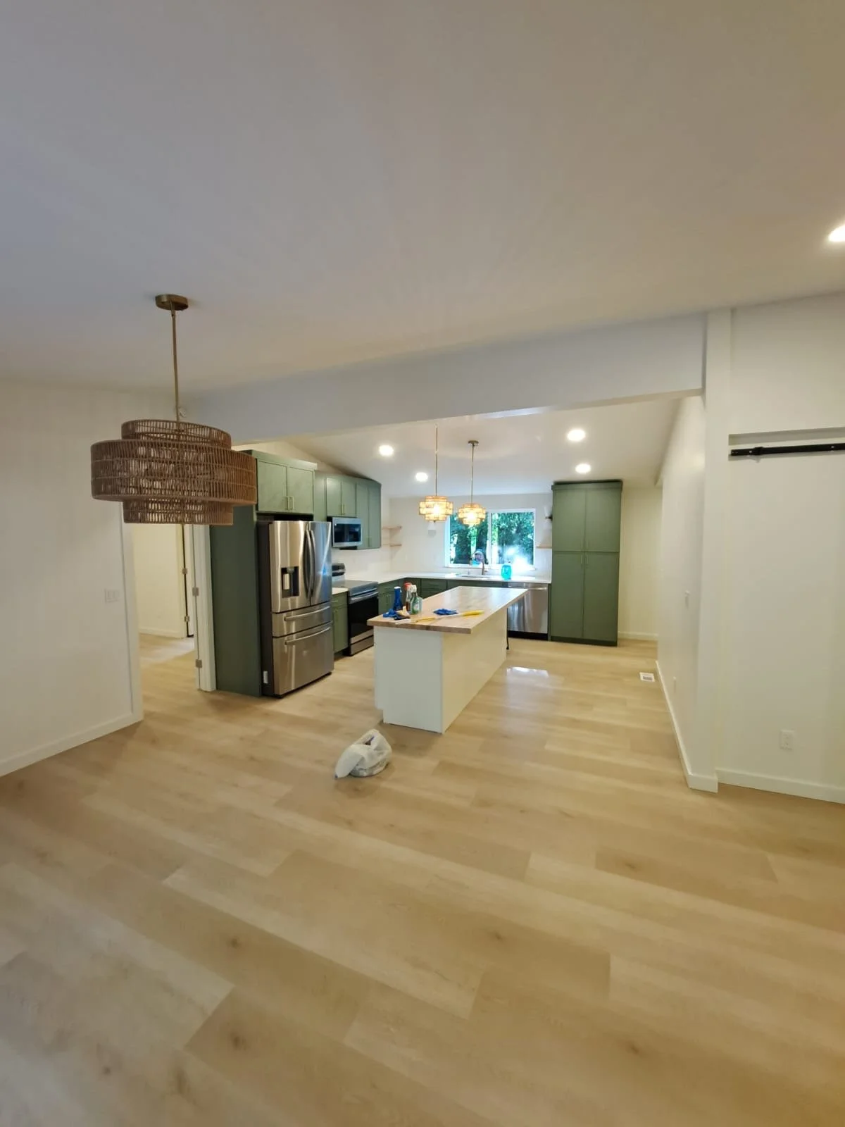 Empty kitchen with light wood flooring, green cabinets, stainless steel appliances, and a kitchen island. There are three pendant lights hanging above the island and two additional ceiling lights. A window provides natural light, and there is a plast