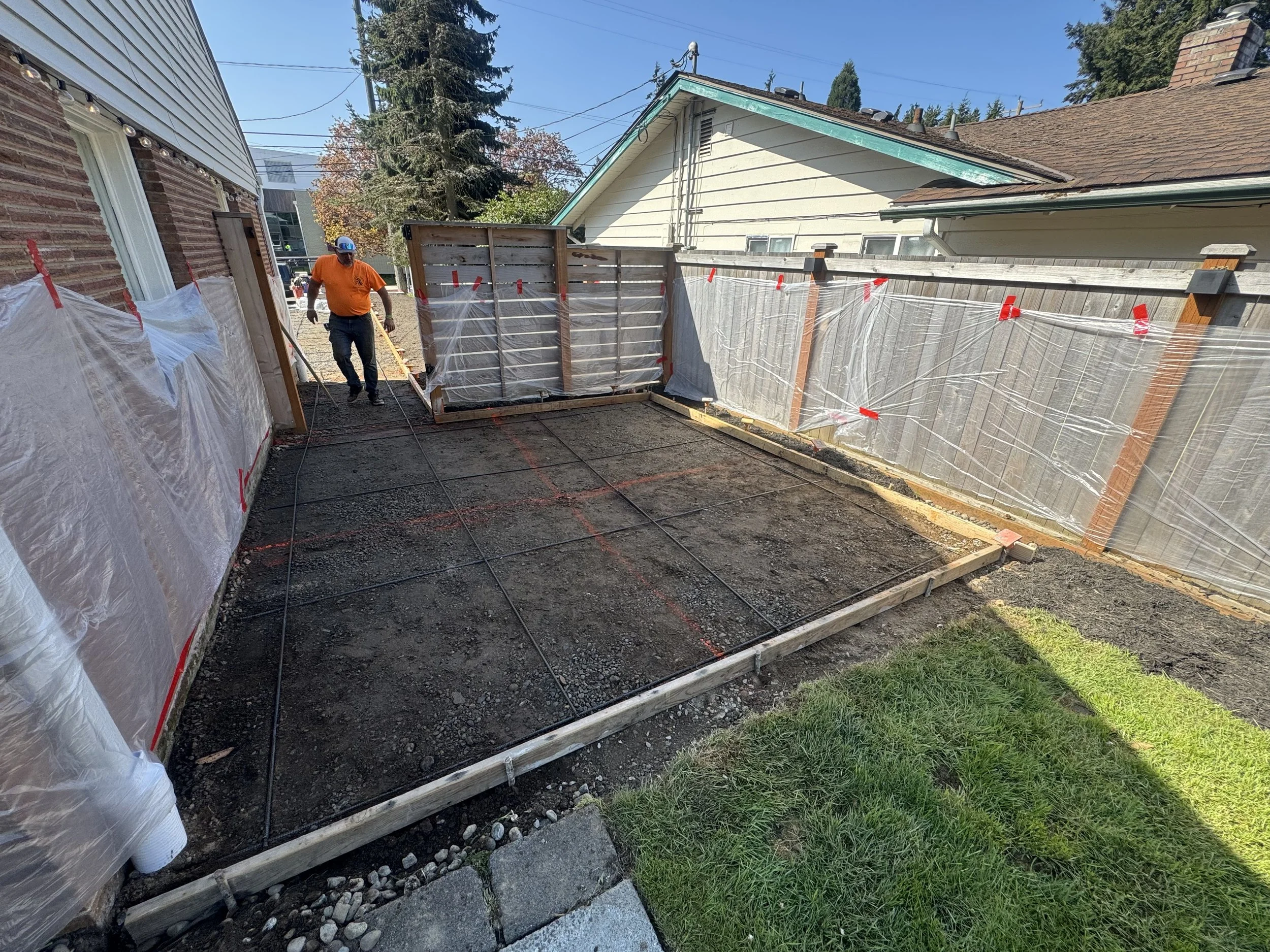A person in an orange shirt and blue hard hat working on a construction site, laying a concrete slab in a small backyard area, with plastic-covered fences and wooden framing.