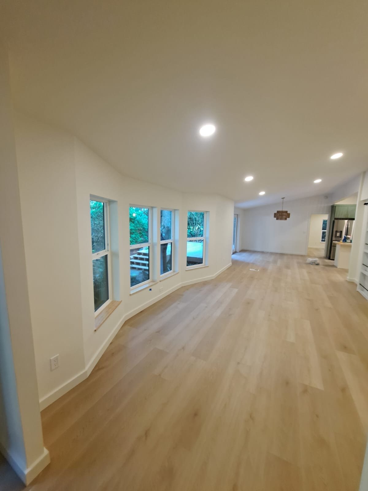 Empty living room with light wooden flooring, white walls, multiple windows, and ceiling lights.