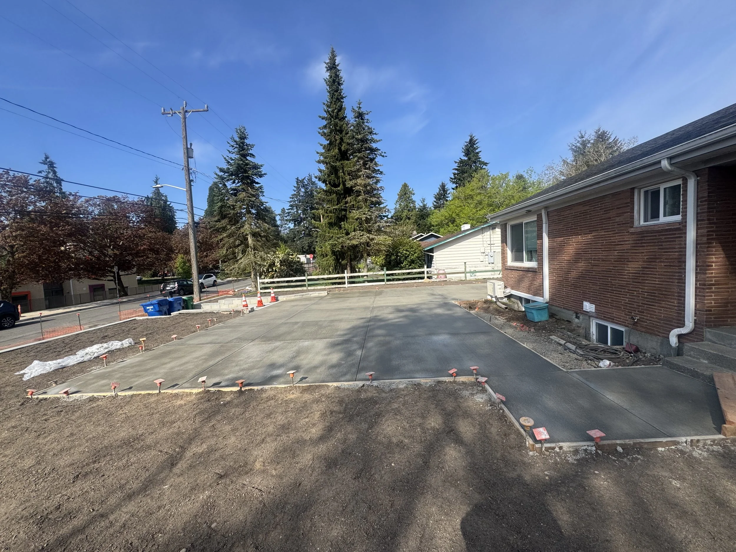 Newly poured concrete driveway with orange markers and a partially finished section. A brick house with white gutters and an air conditioning unit is visible on the right. Trees and utility poles are in the background on a sunny day.