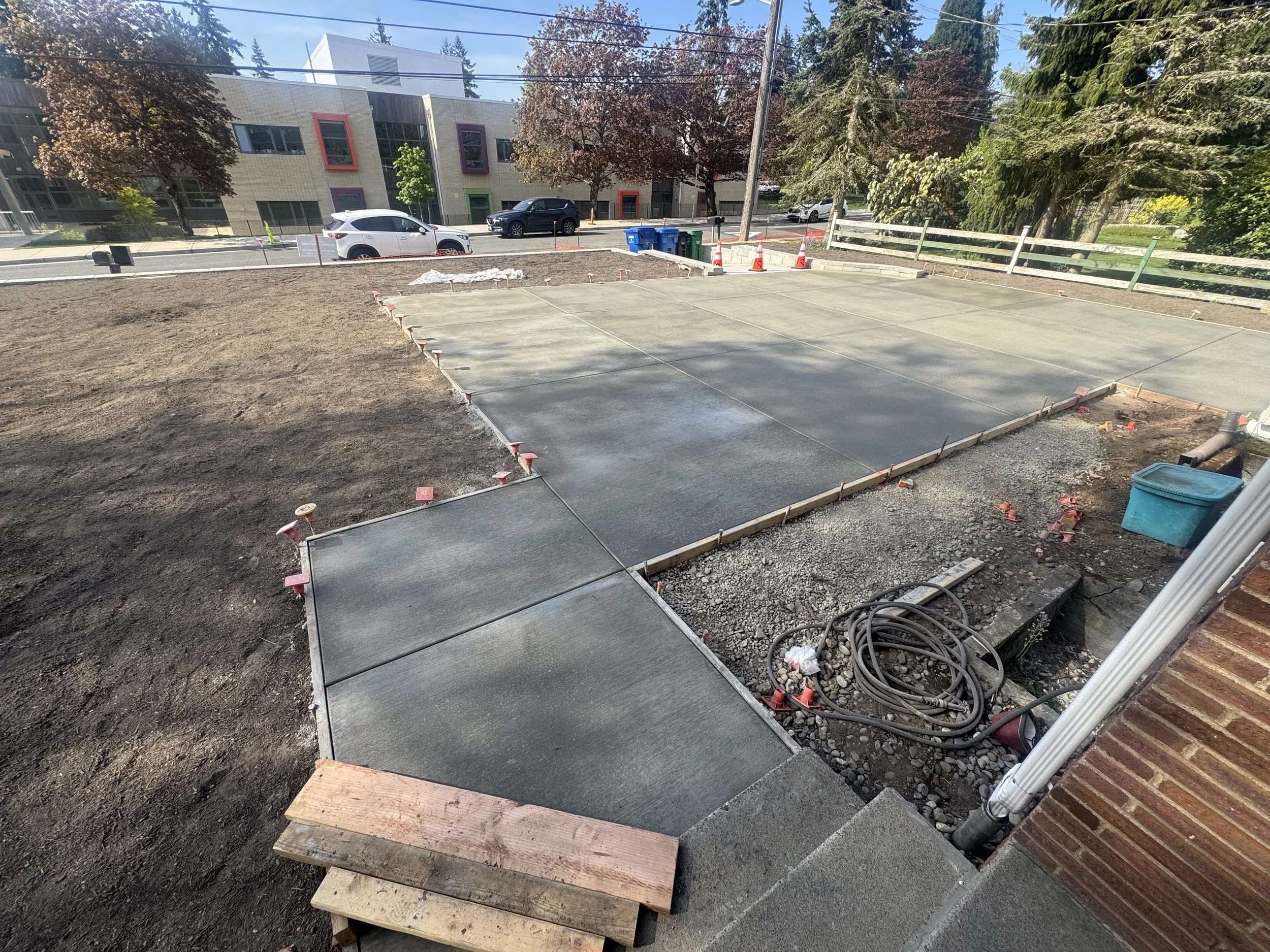 Newly poured concrete sidewalk section next to a building, with construction tools and gravel nearby, and a parking lot and street with cars in the background.