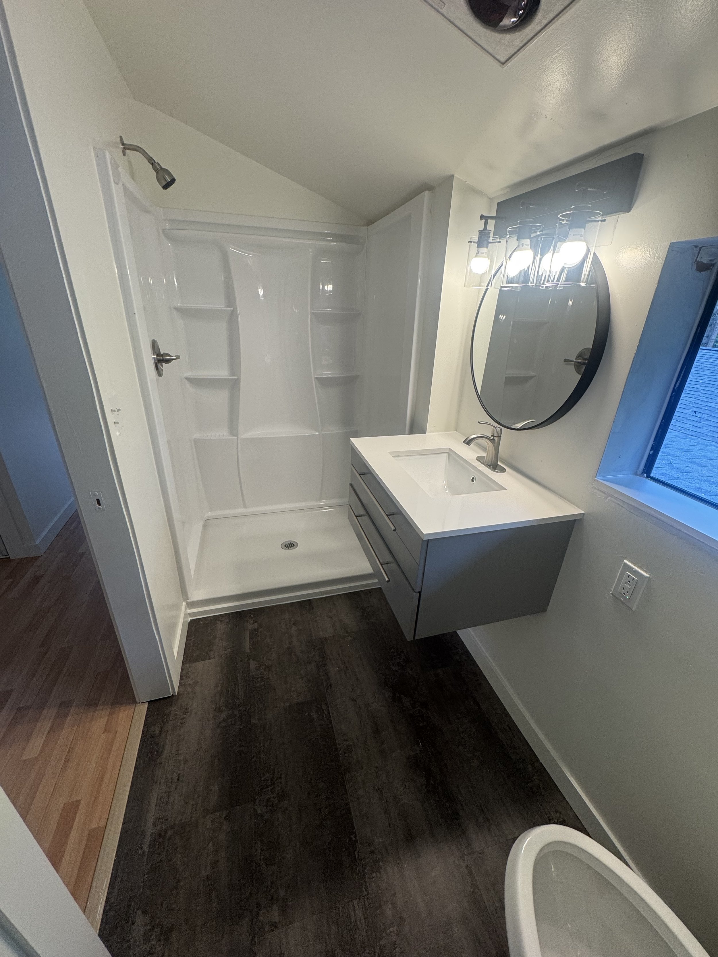 Bathroom with a walk-in shower, vanity with a sink, round mirror, modern lighting fixture, and a window.