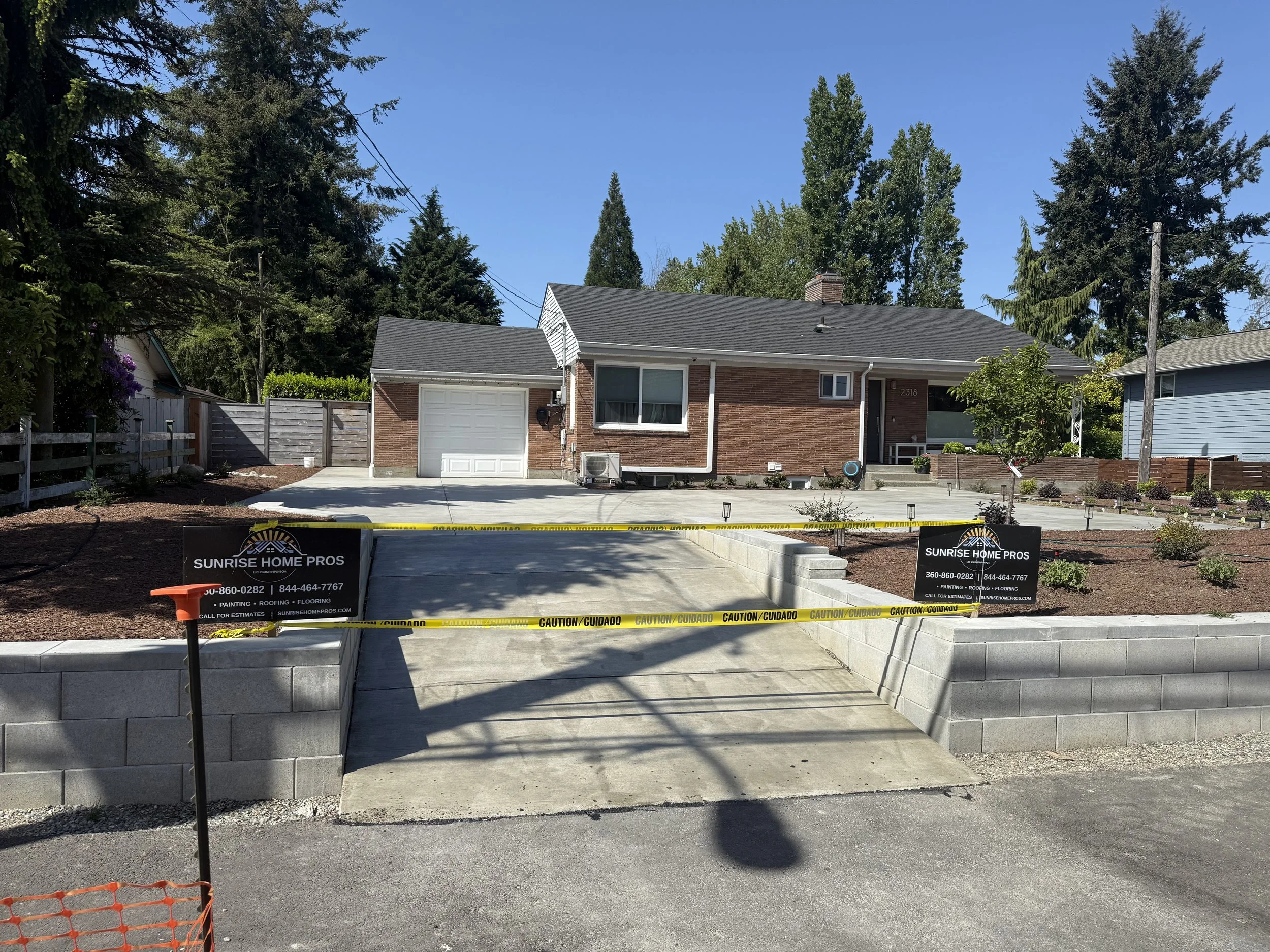 A newly paved driveway in front of a brick house, surrounded by a low concrete block wall and caution tape, with signs for Sunrise Home Pros for roofing, painting, and flooring services.