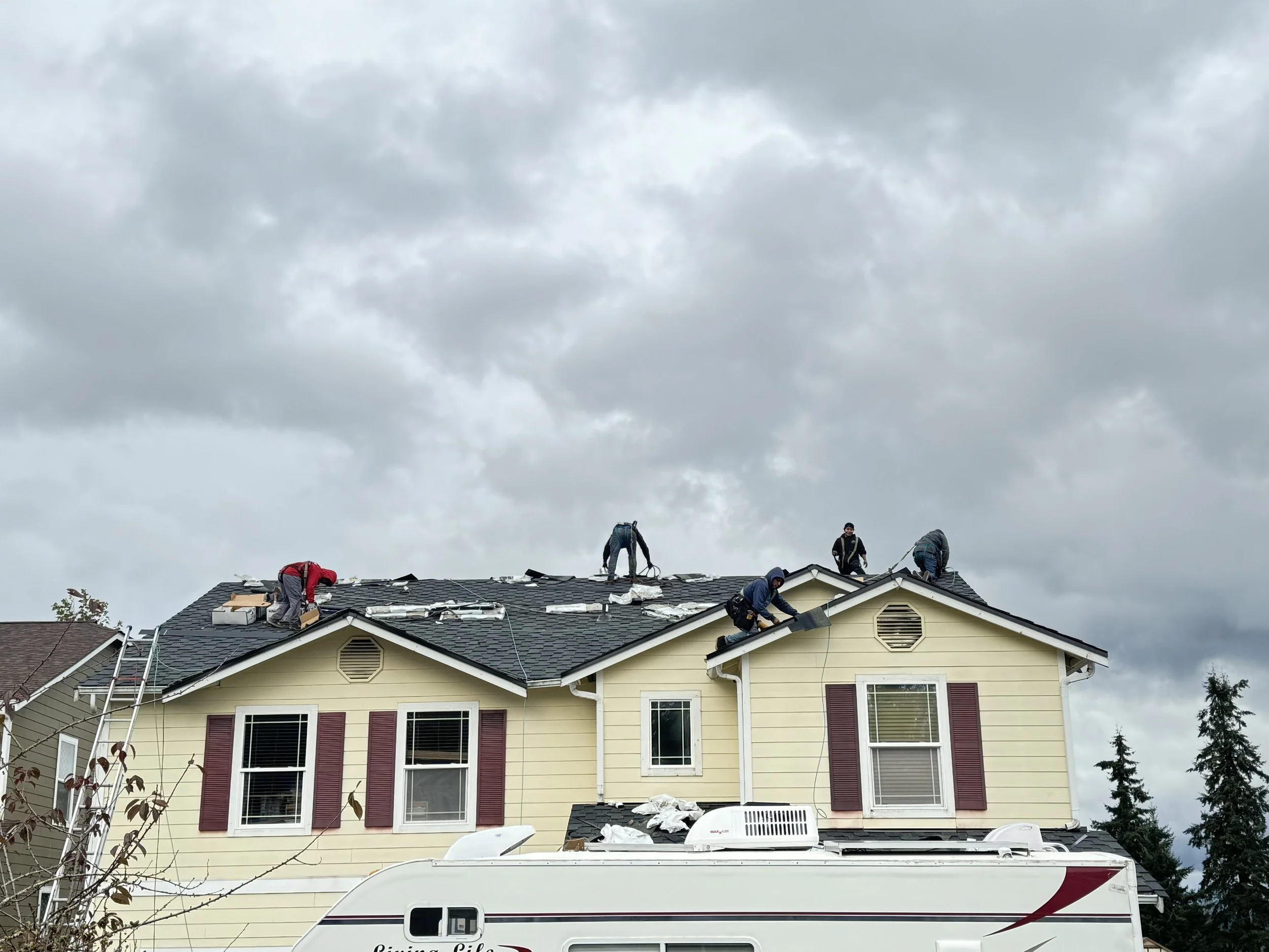 Roofers working on a house roof during cloudy weather, with some workers kneeling and others standing on the roof, and a camper trailer parked in front.