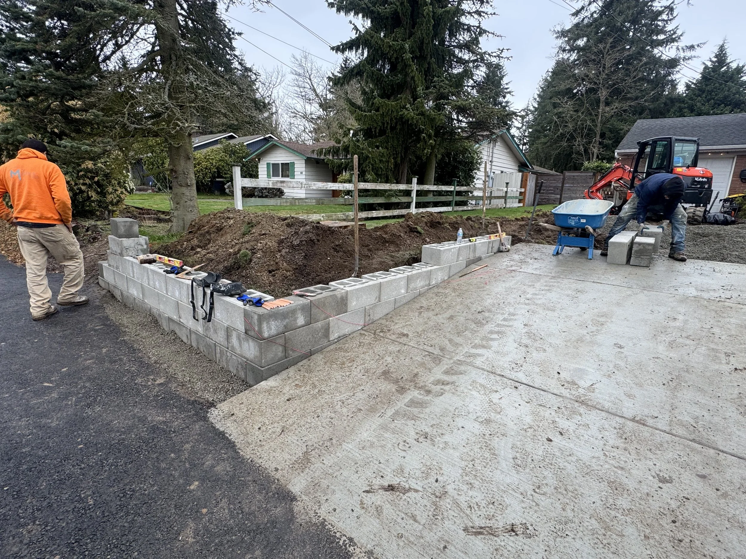 Construction workers building a cinder block wall on a residential driveway in a suburban neighborhood with trees and houses in the background.