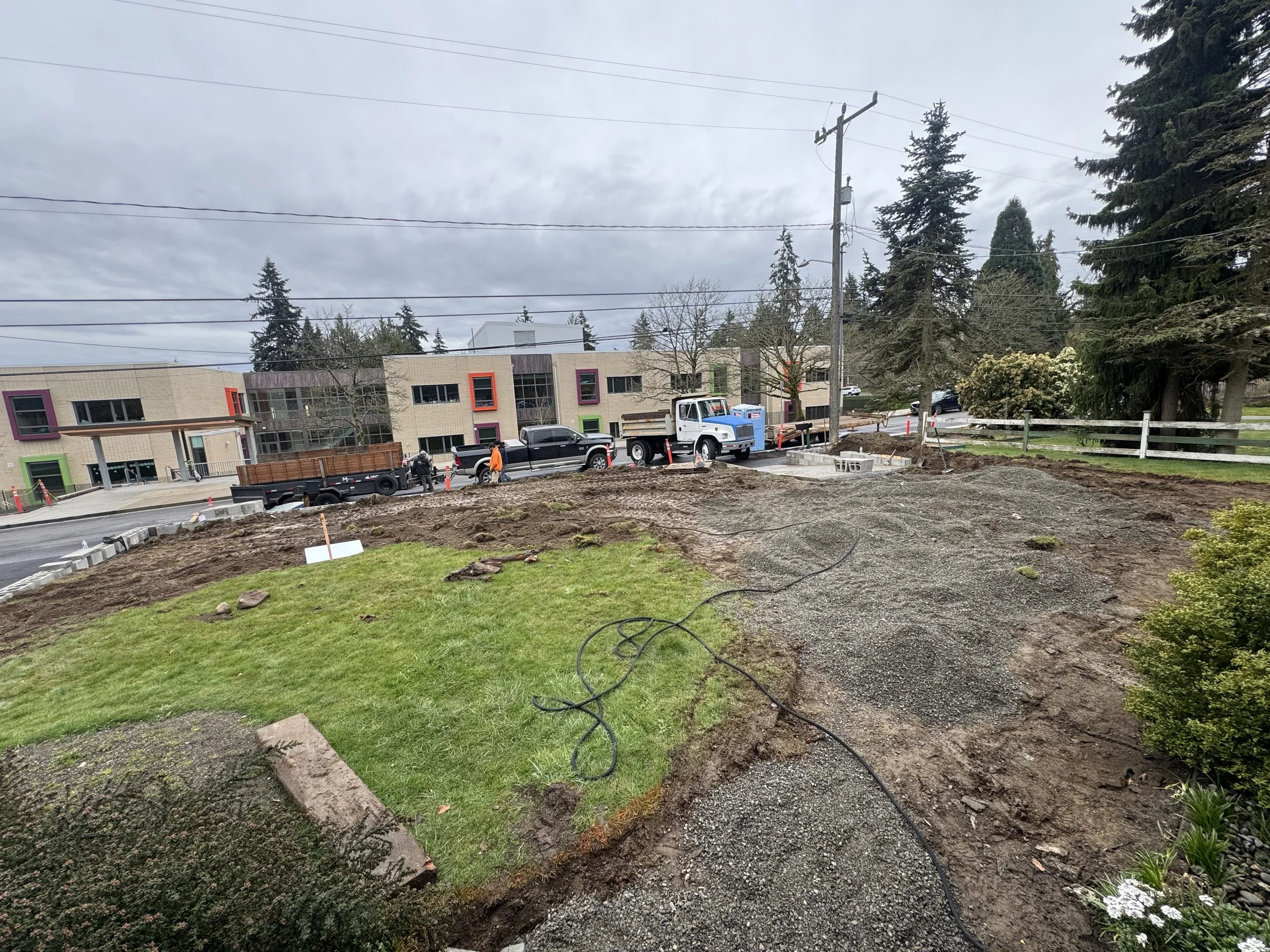 Construction site with trucks and workers near a modern building, with dirt and gravel areas and trees in the background.