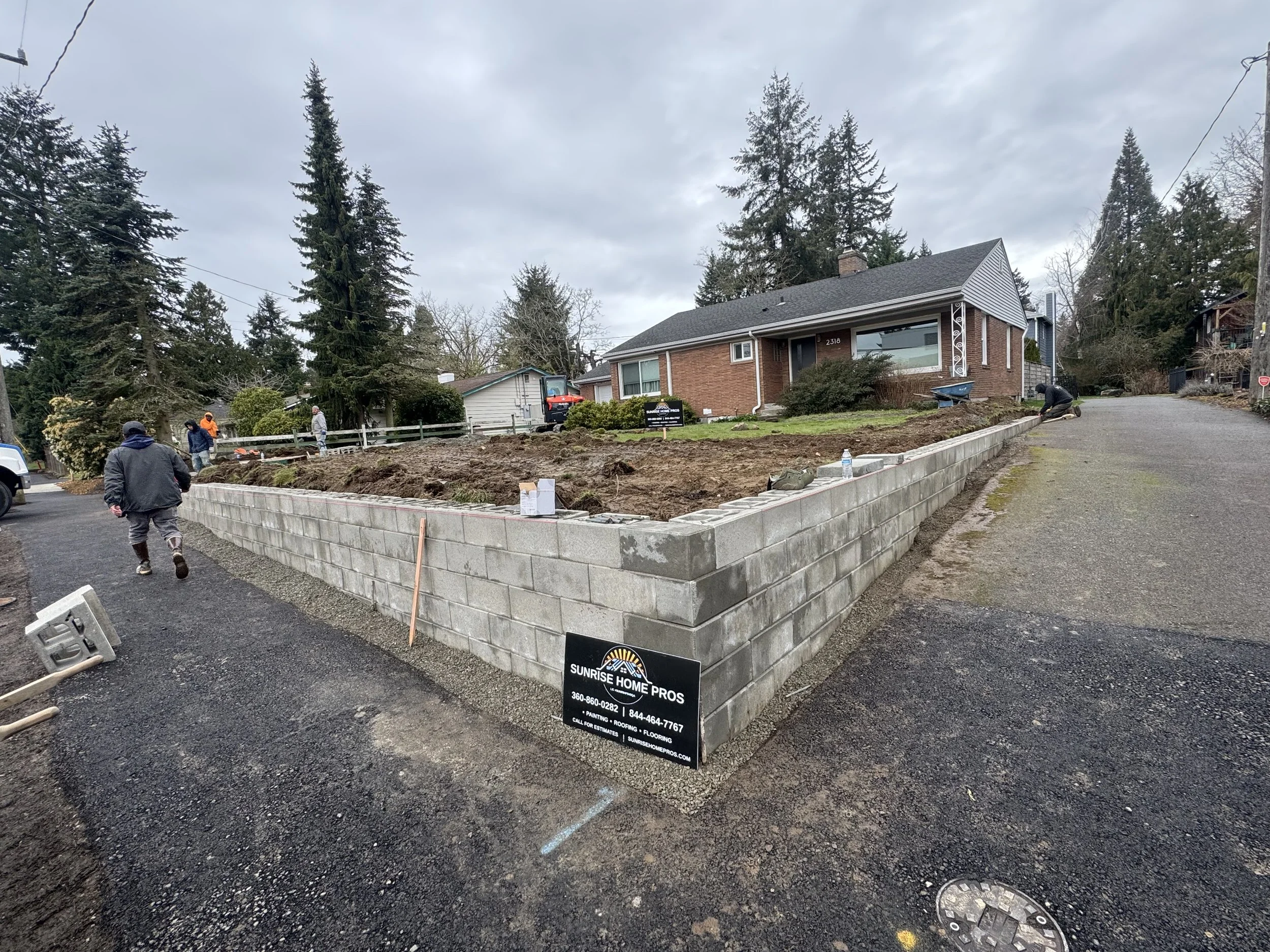 Construction workers building a brick garden wall in front of a house, with trees and cloudy sky in the background.